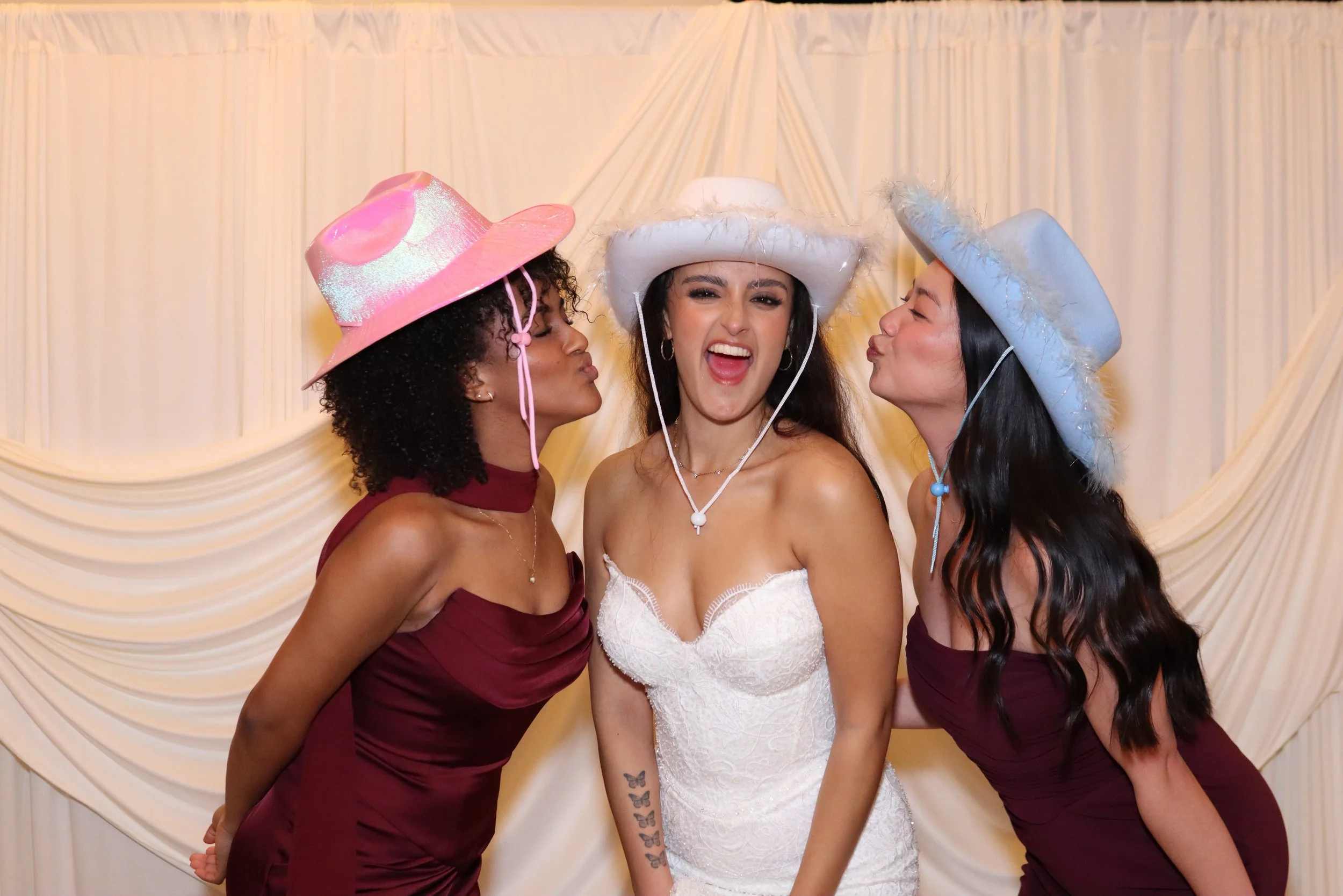 Three women in party hats and dresses smiling and kissing at a celebration or wedding reception.