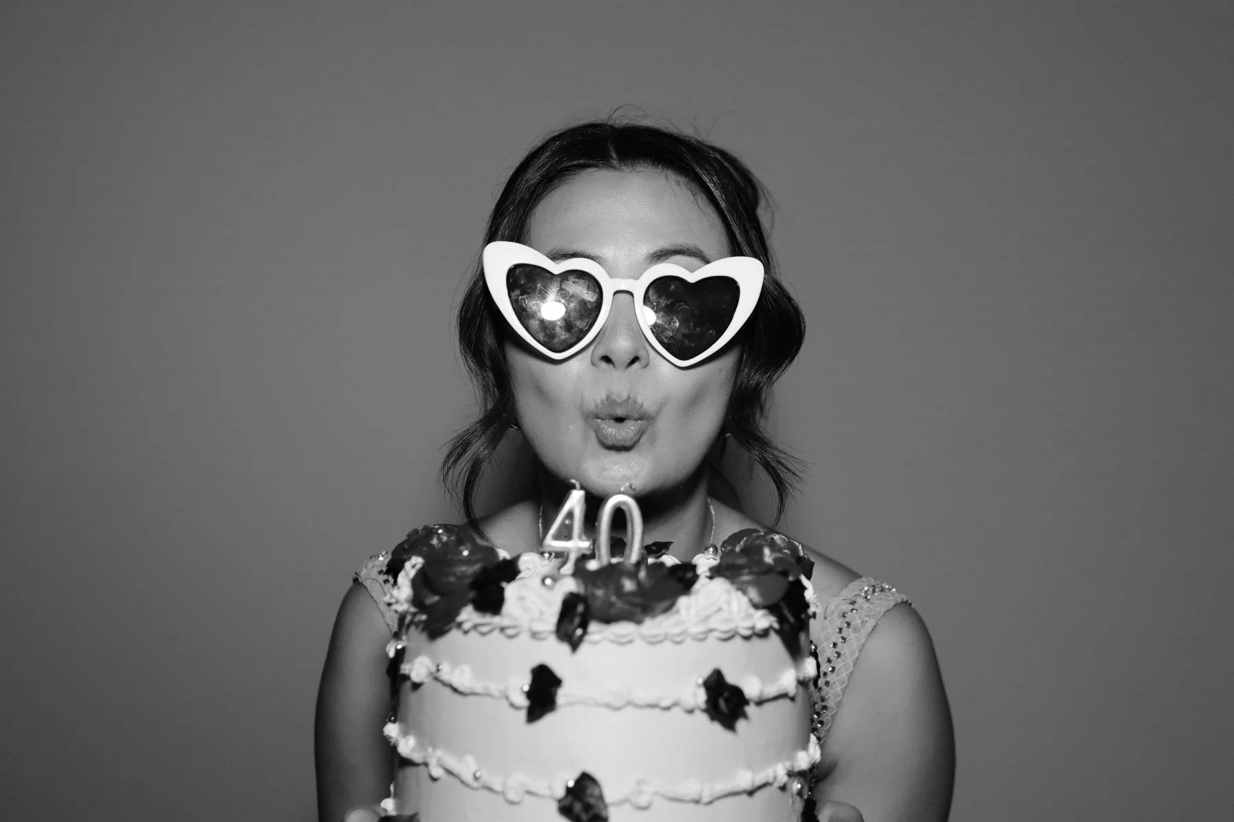 A woman with shoulder-length hair wearing heart-shaped sunglasses, holding a birthday cake with the number 40 on top, and blowing a kiss.