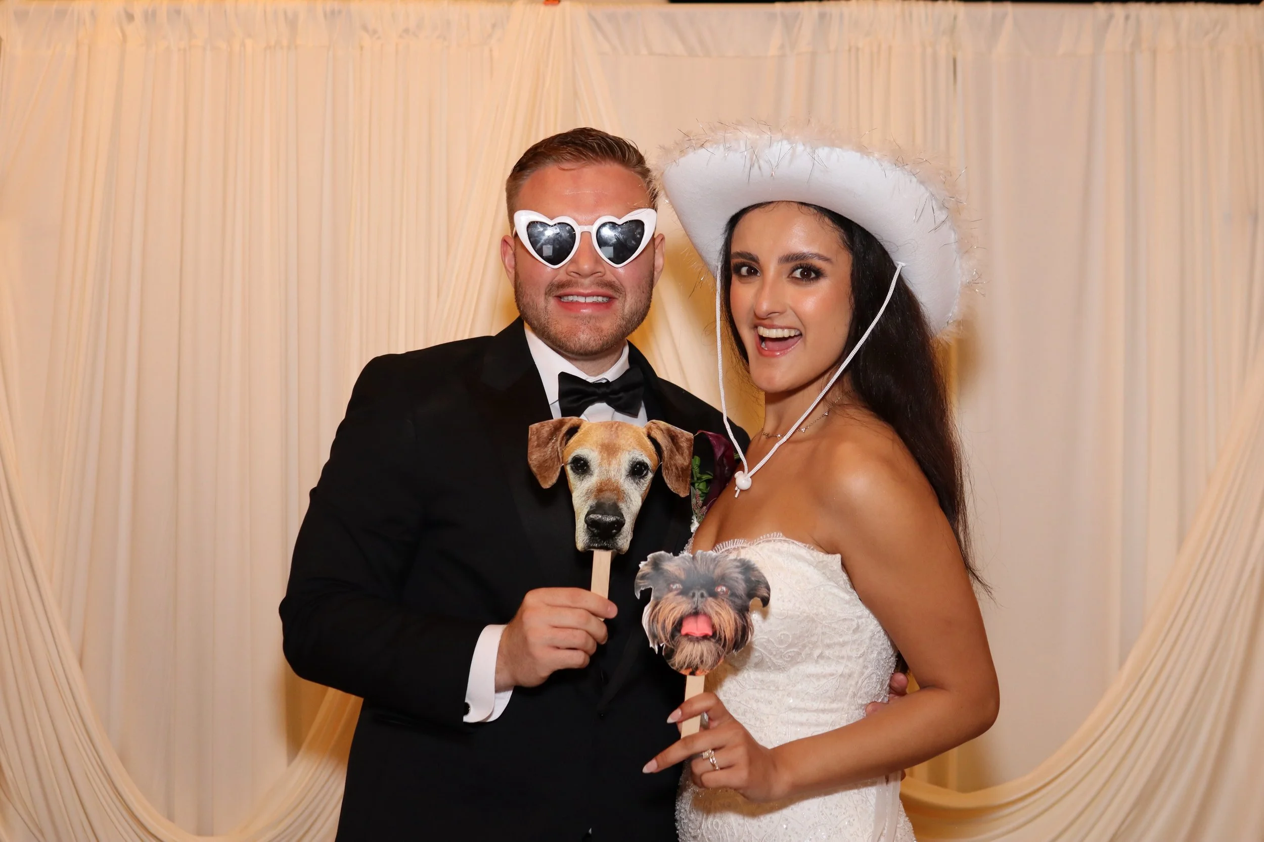 Couple at a wedding or event, wearing fun accessories like sunglasses and a hat, holding dog-themed photo props, standing in front of a cream-colored draped backdrop.