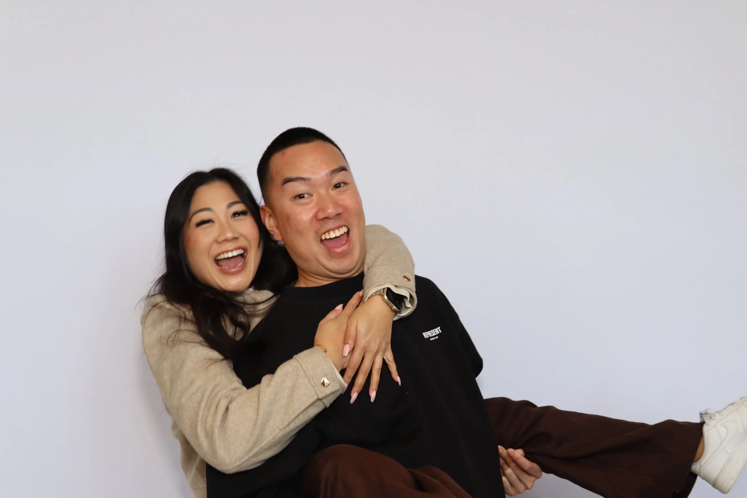 A smiling woman playfully piggybacking a smiling man against a plain white wall.