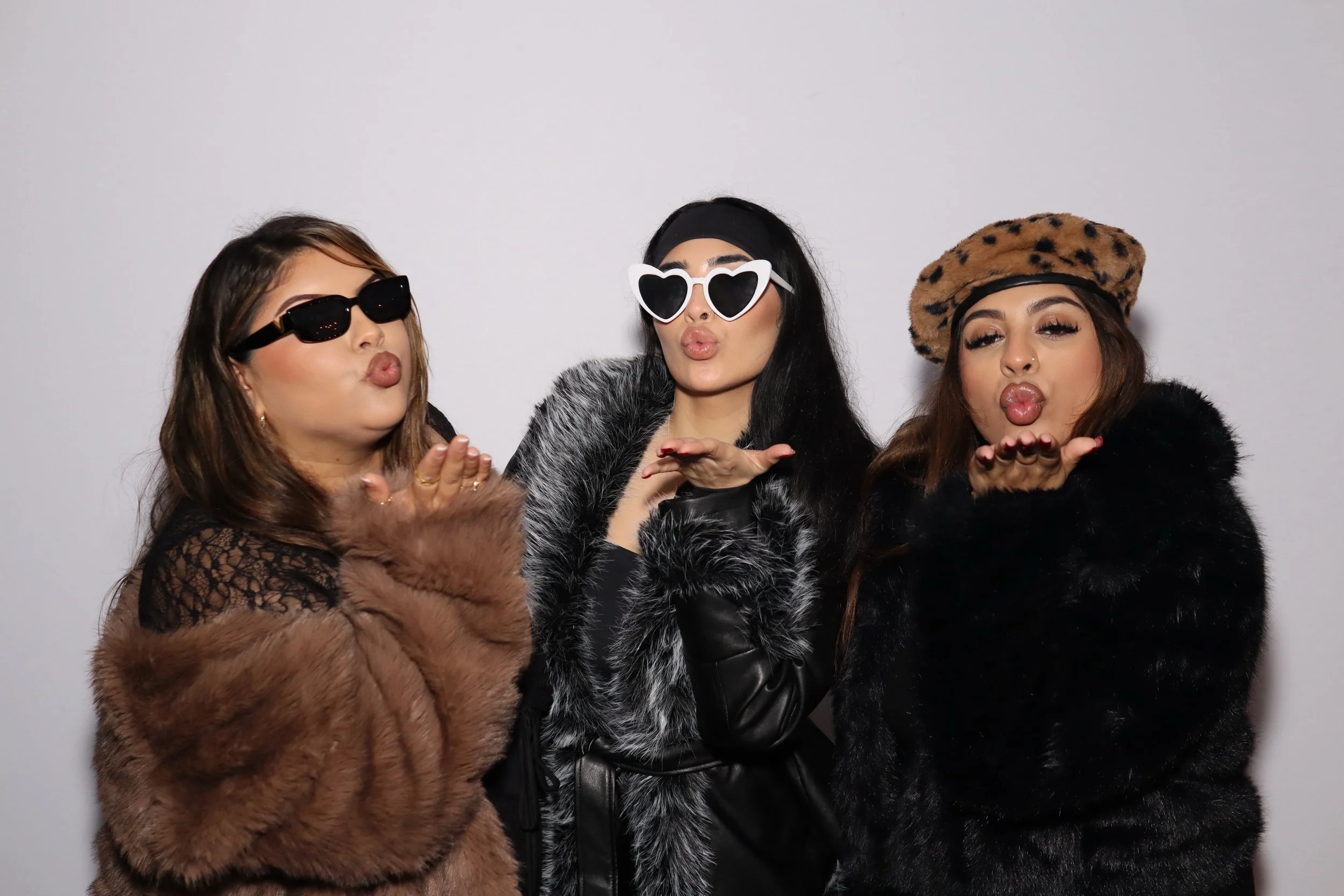 Three women wearing fashionable clothing and sunglasses, blowing kisses toward the camera, standing against a plain white background.