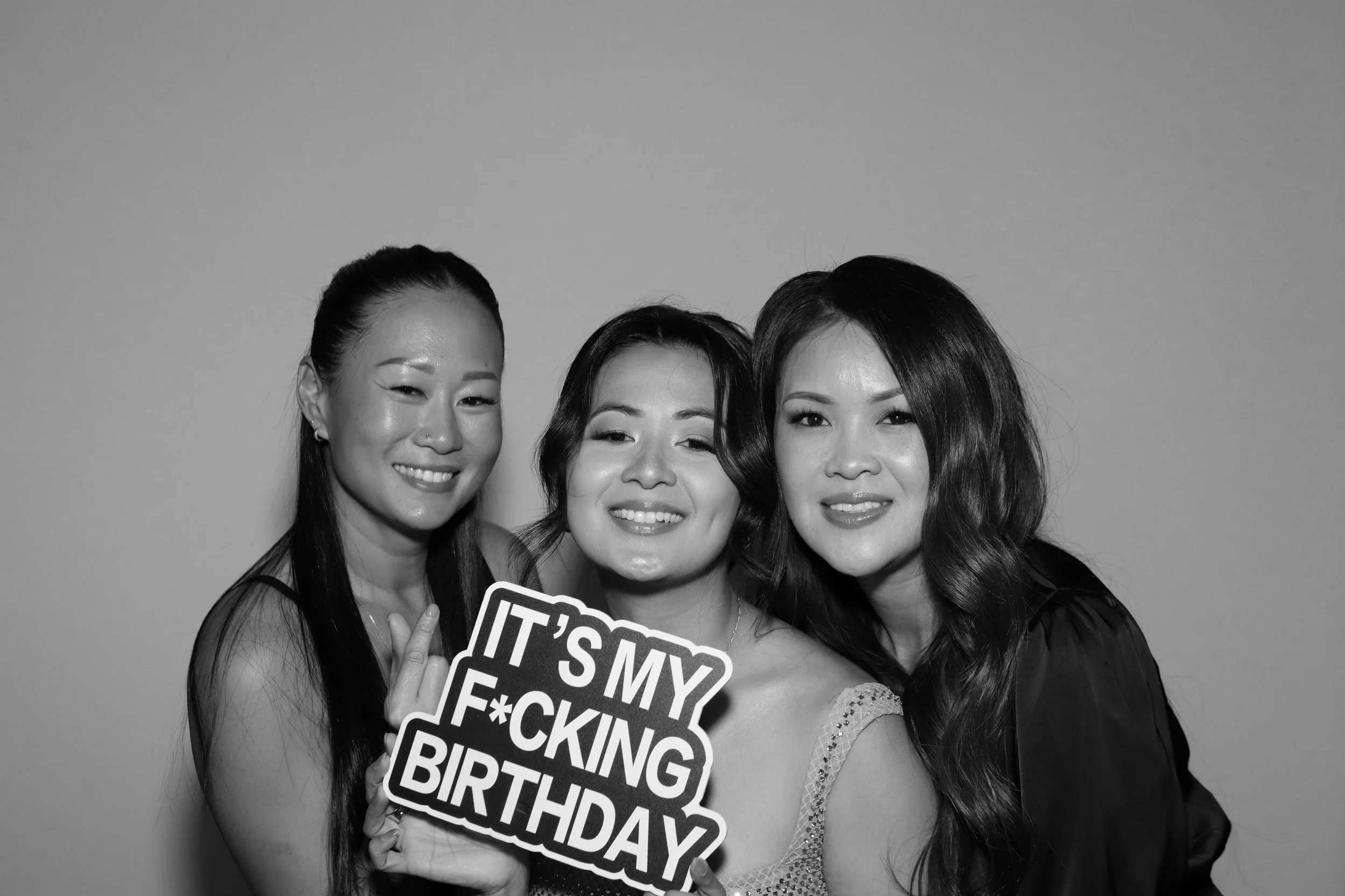 Three women smiling at a birthday celebration, holding a sign that says "It's my f*cking birthday" in black and white.