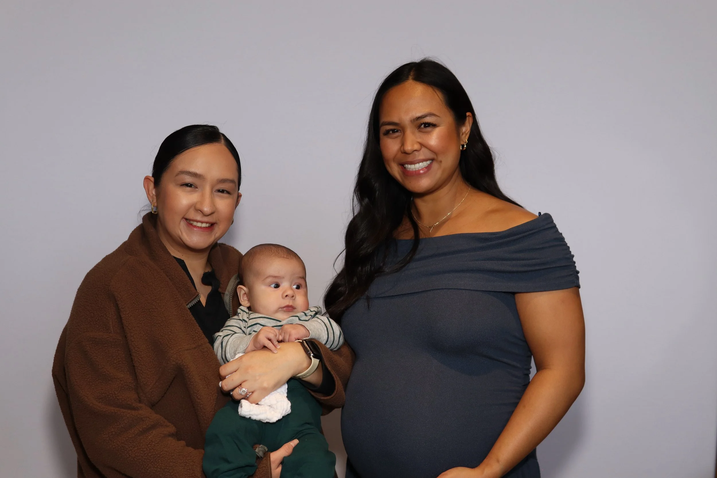 Three people, two women and a baby, standing against a plain background smiling for the camera.