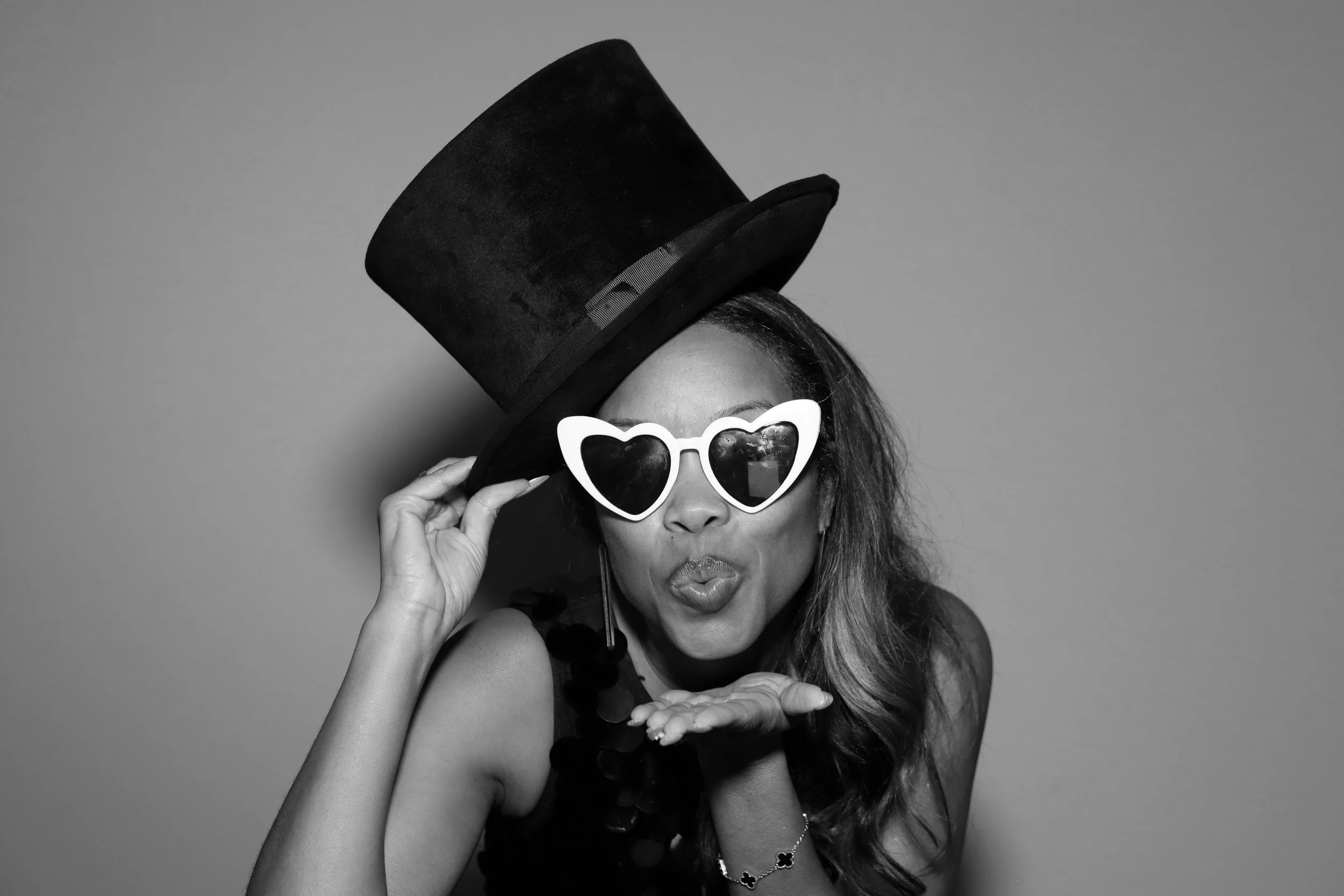 Woman dressed in fashionable accessories, wearing heart-shaped sunglasses, and a large top hat. She is blowing a kiss and holding the brim of her hat.