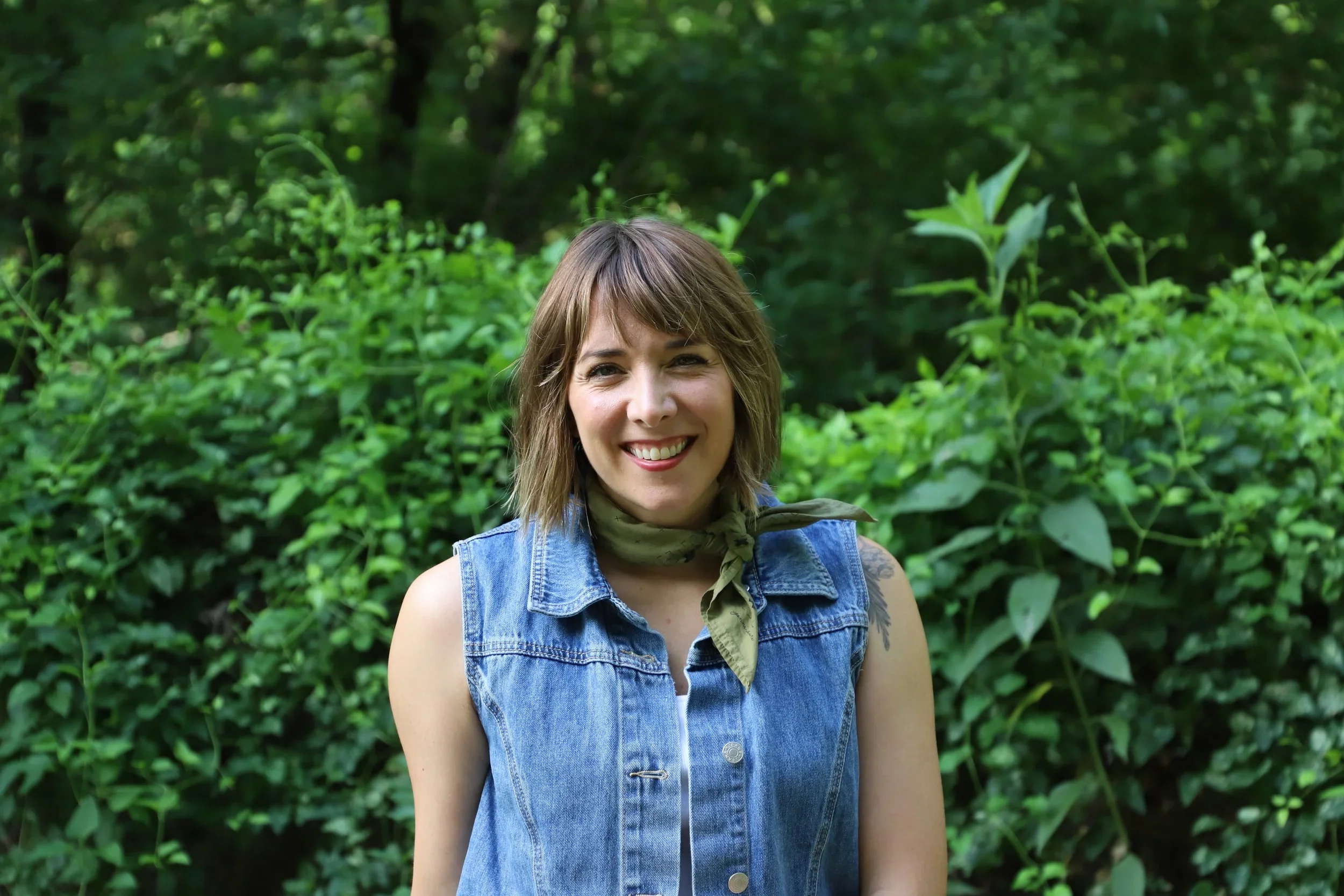 A young woman with brown hair and a green scarf smiling in front of lush green foliage.