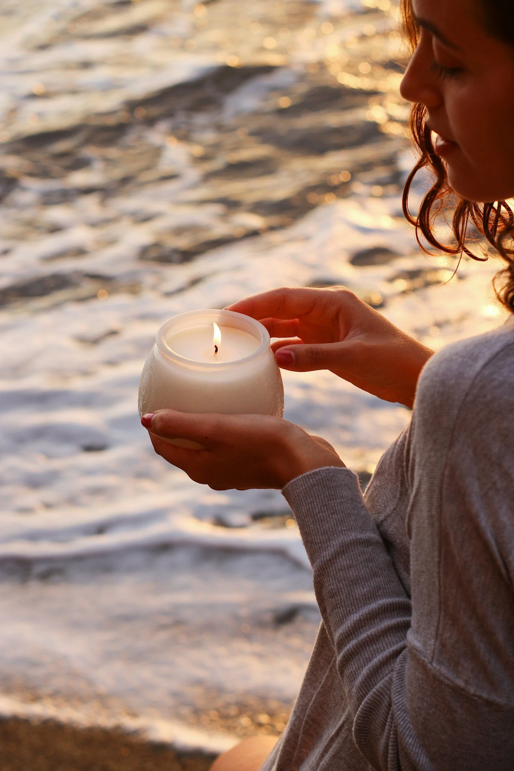 A woman holding a lit candle in a glass container on a beach during sunset.