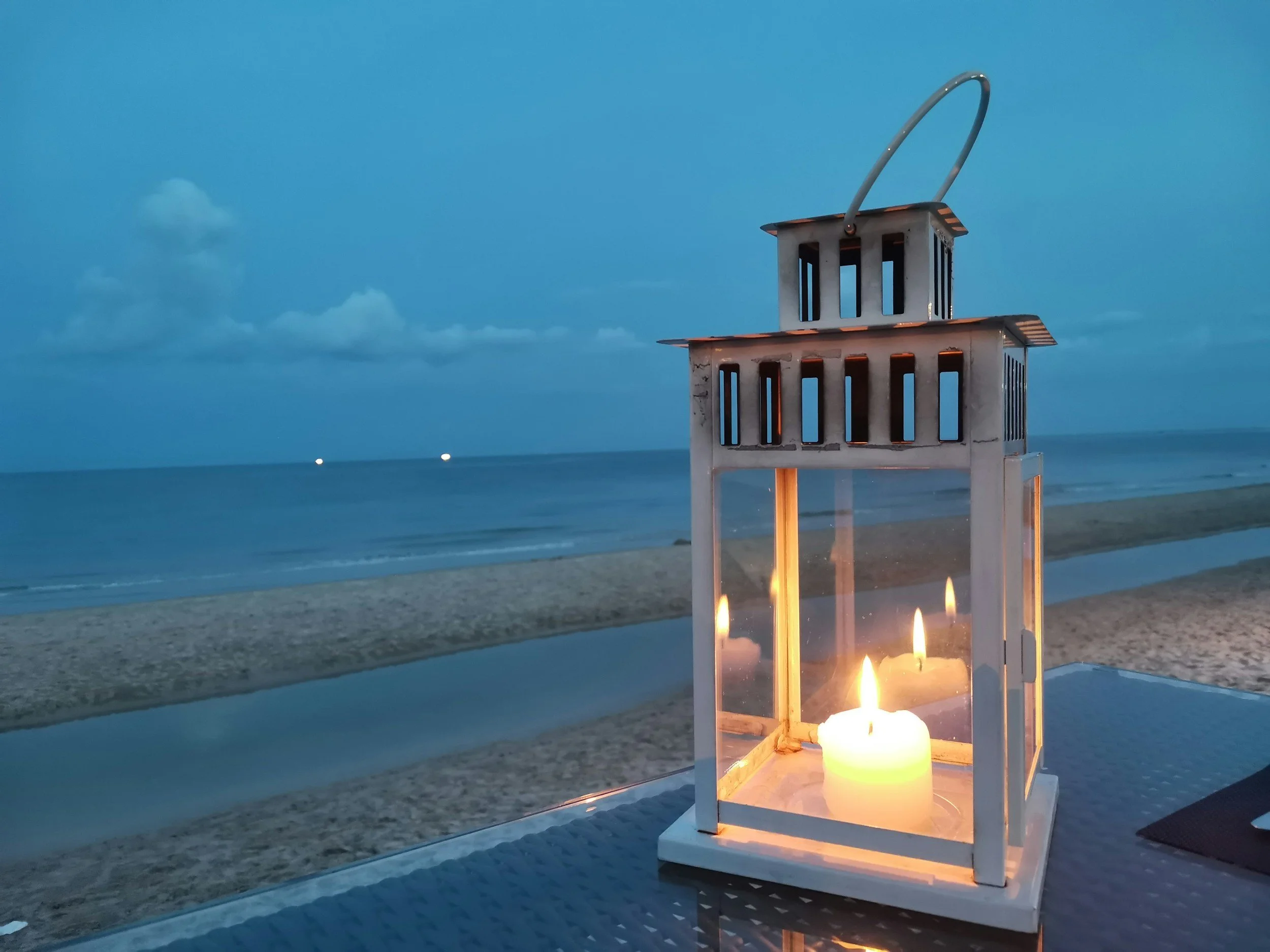 A white lantern with a flickering candle inside, placed on a table on a beach at dusk, with an ocean view and a cloudy sky in the background.