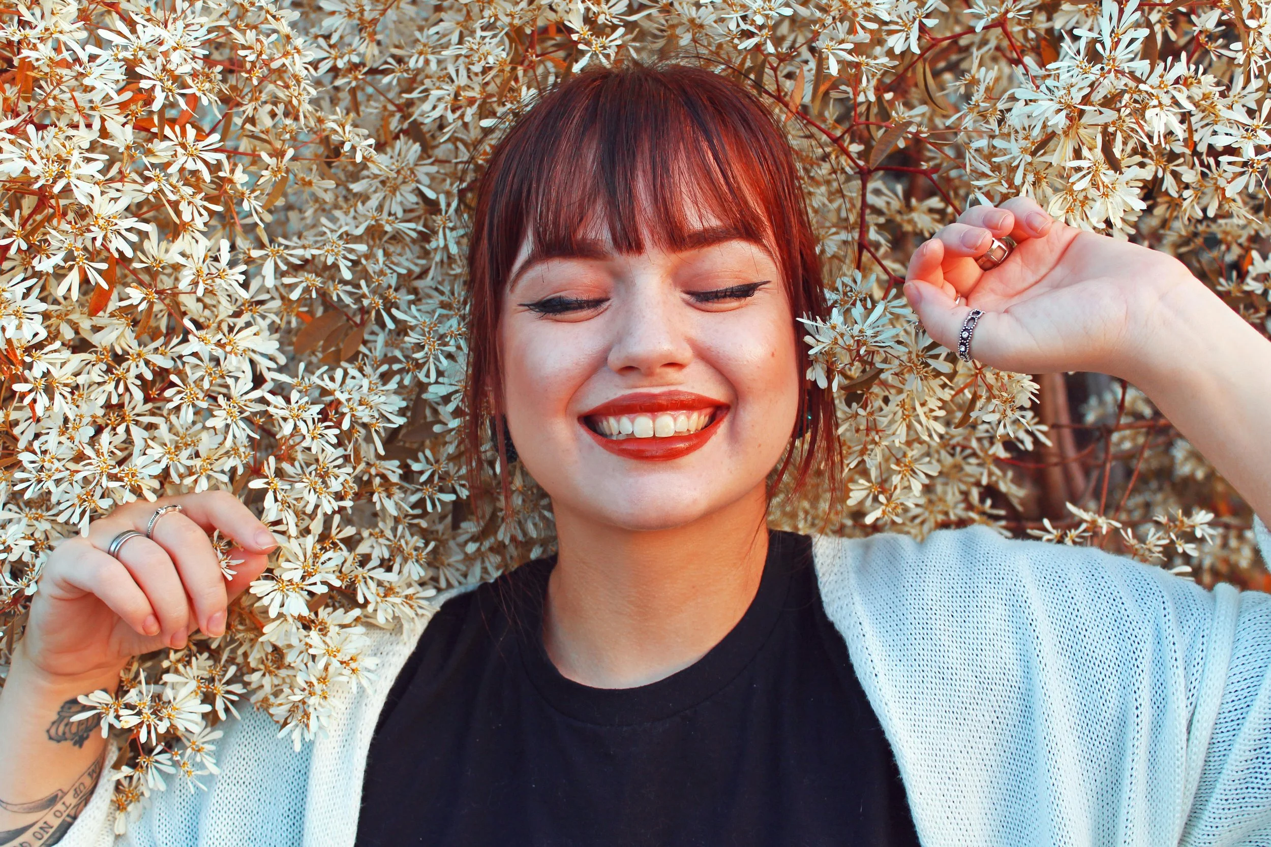 A woman with red hair and bangs smiling with eyes closed, surrounded by white flowering branches, wearing a black shirt and a white cardigan, with rings on her fingers.