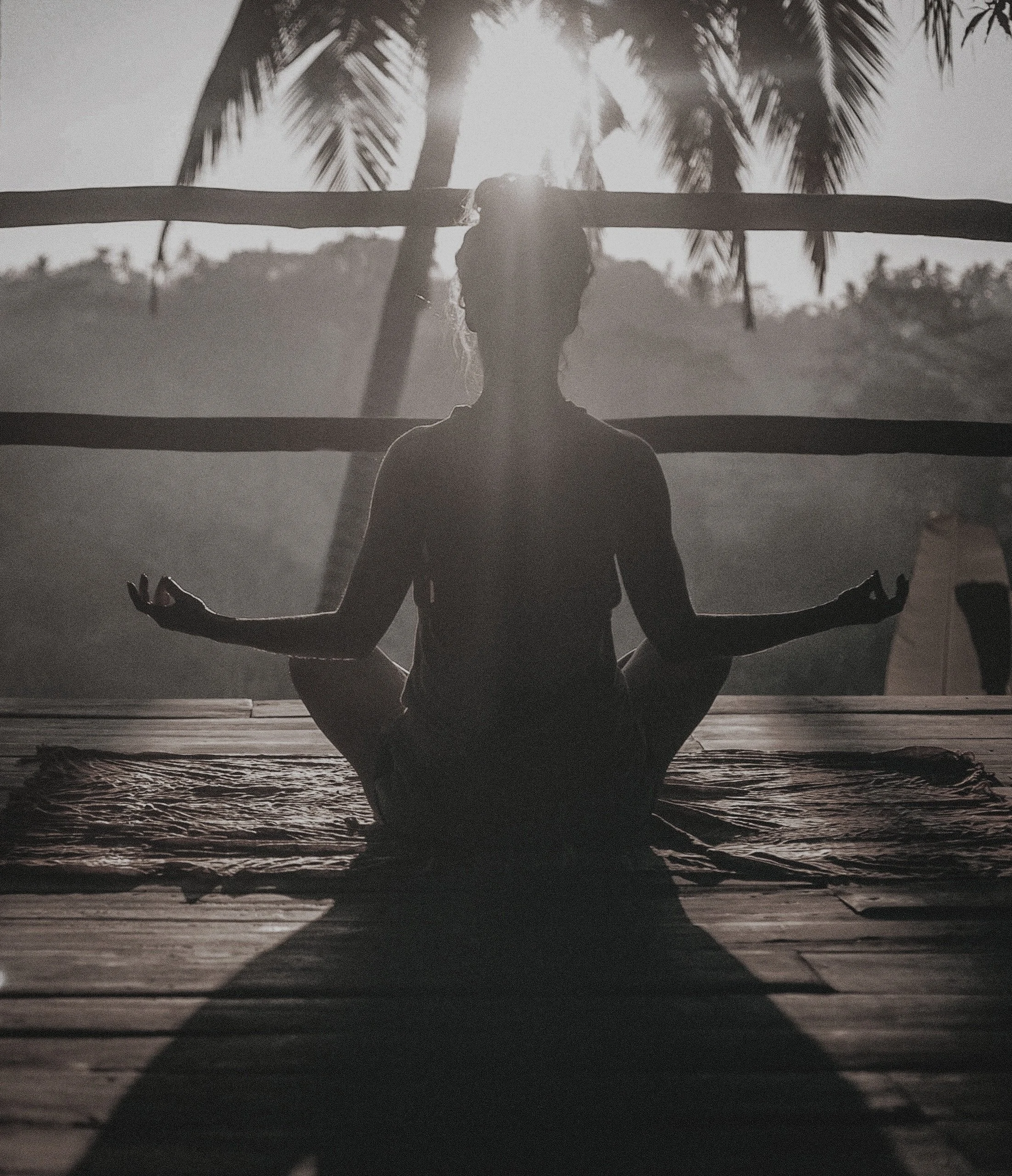 A person practicing meditation or yoga outdoors in a seated cross-legged position on a mat with arms extended, hands in a mudra gesture, silhouetted against the sunlight coming through palm trees in the background.