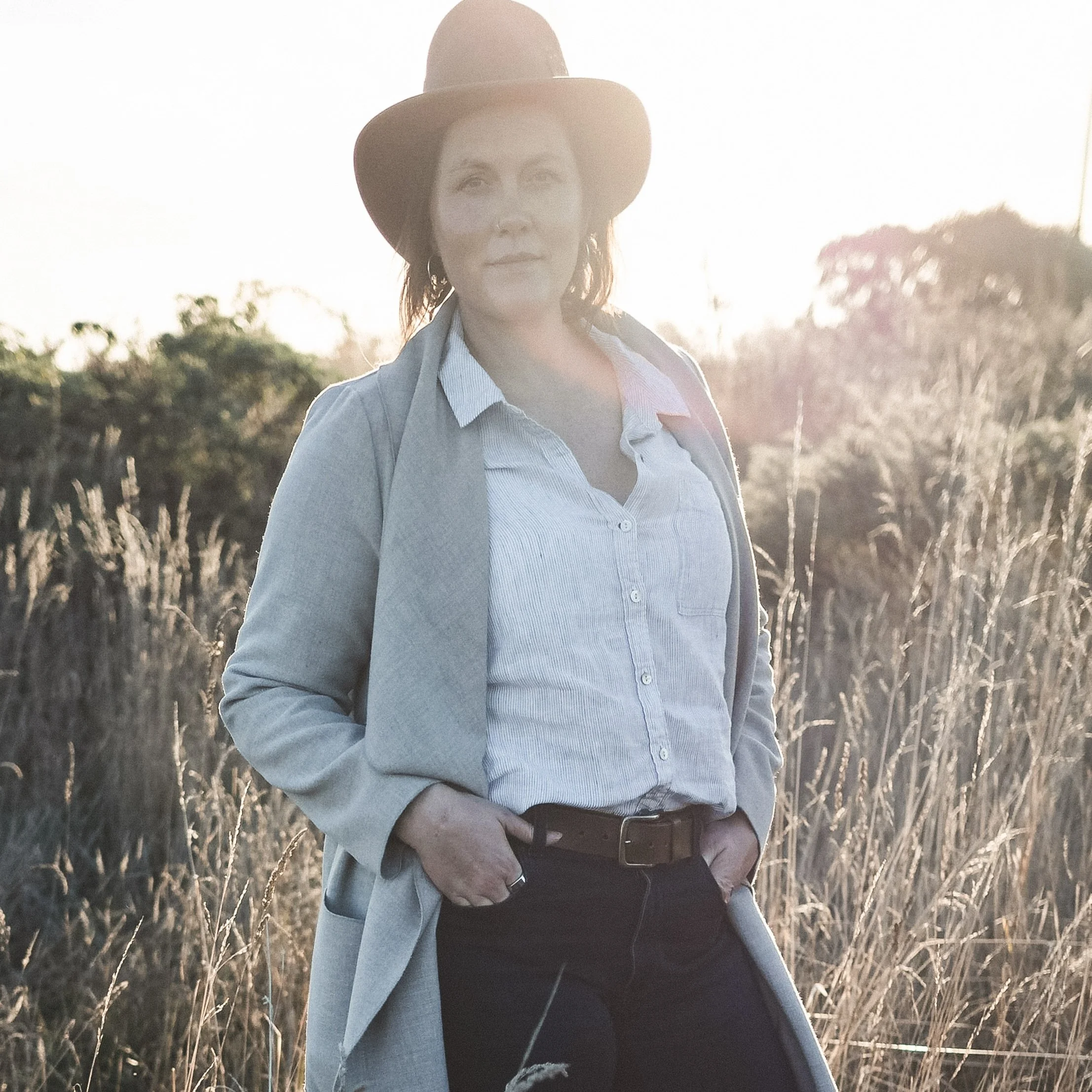A woman with a hat outdoors in a field, wearing a light-colored shirt, gray coat, and black pants, with her hands in her pockets during sunset.