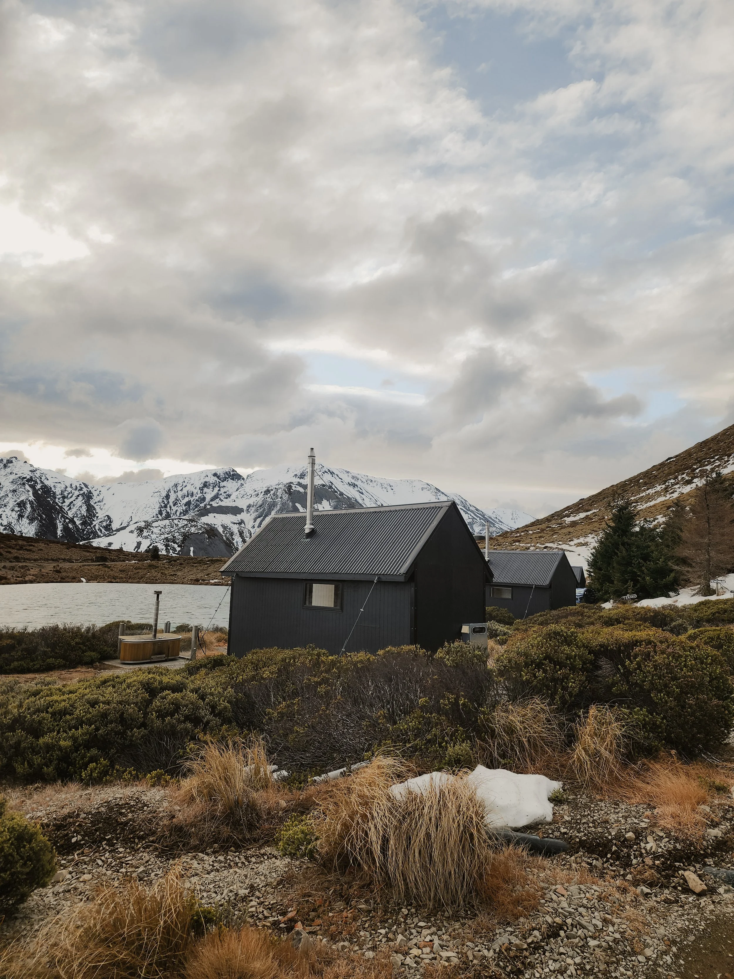 Black wooden cottages with snow-capped mountains in the background, cloudy sky, and dry vegetation in foreground.
