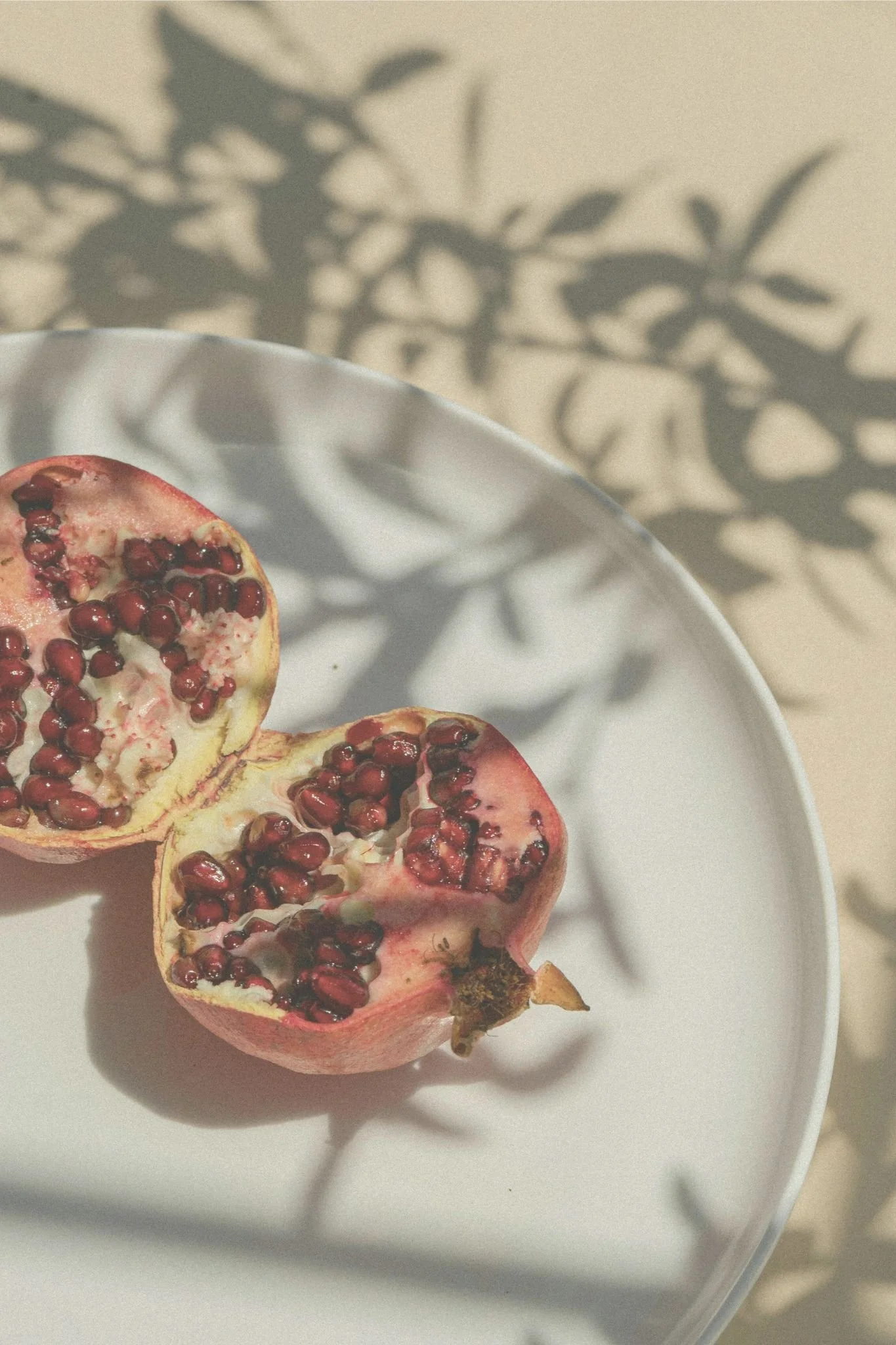 A halved pomegranate fruit with red seeds on a white dish, casting shadow on a beige surface.