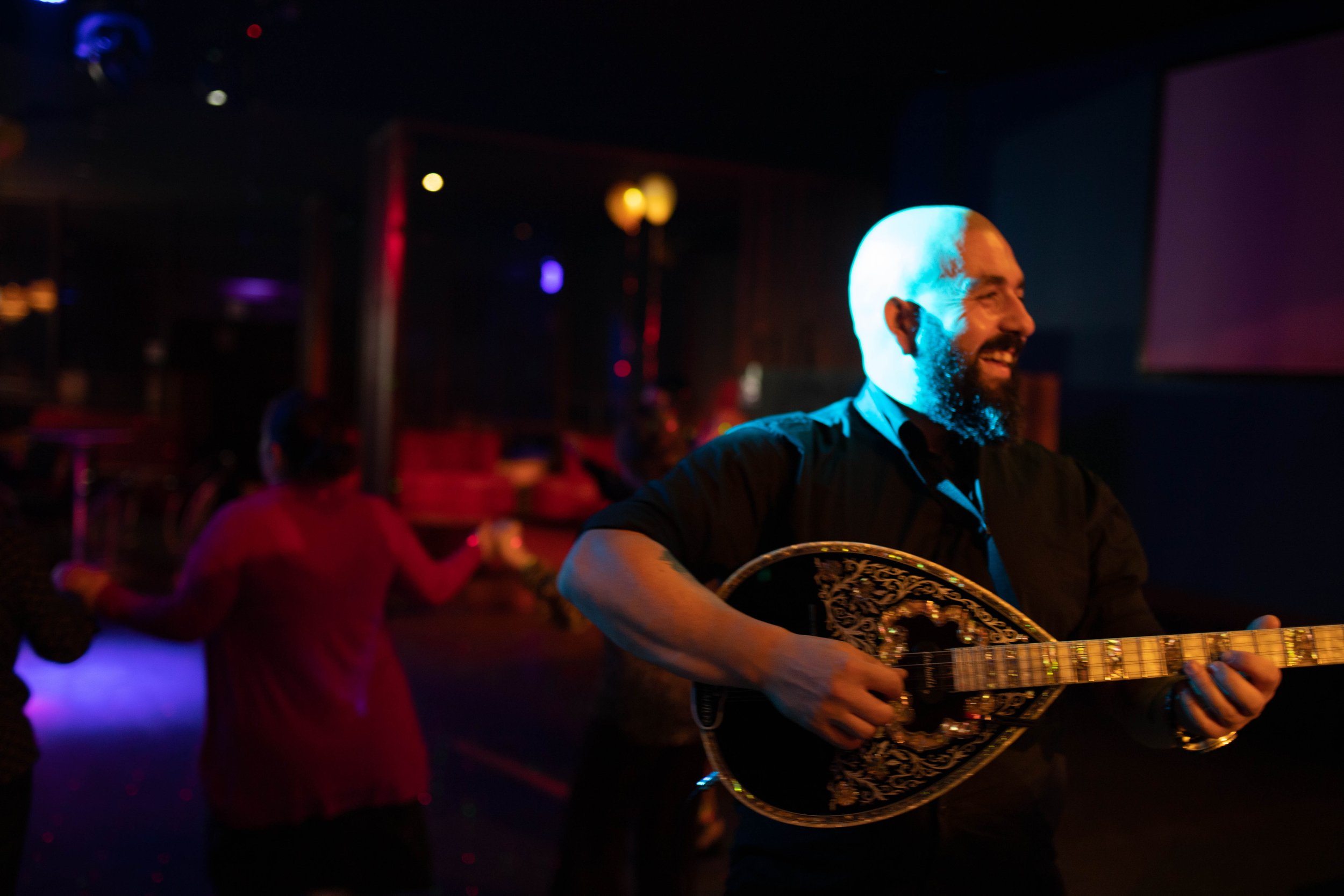 A man with a bald head and beard, smiling and playing a decorated string instrument in a dimly lit venue with colorful lights, while people dance in the background.