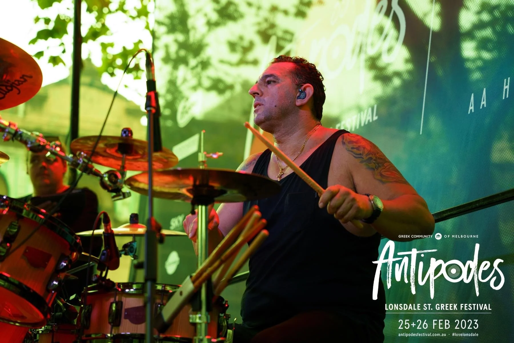 A man playing the drums at the Lonsdale St. Greek Festival, February 25-26, 2023, with green trees and festival logo in the background.