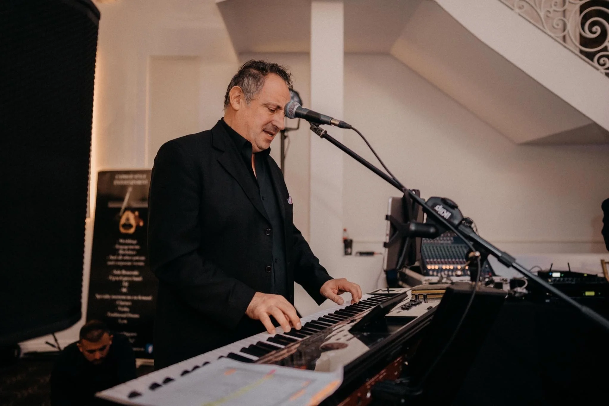 A man in a black suit playing a keyboard at a music event, with a microphone in front of him and audio equipment visible on the side.