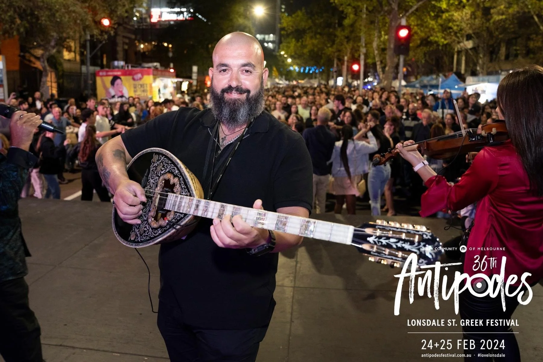 A man with a beard playing a bouzouki at Lonsdale St. Greek Festival, with a crowd of people in the background, at night.