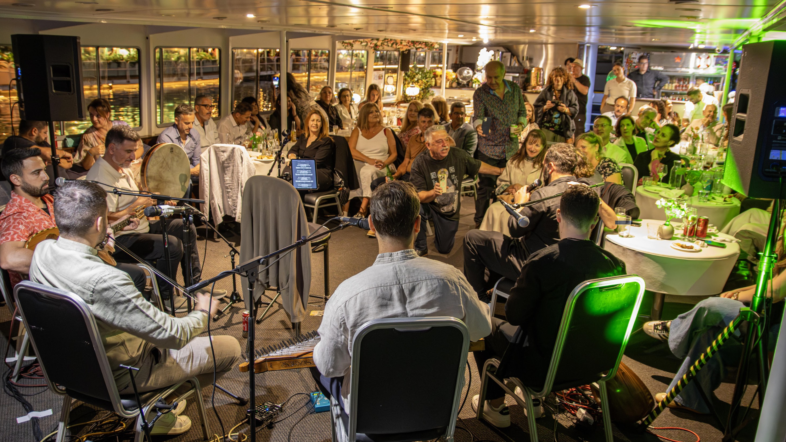 Indoor gathering with musicians performing and audience enjoying live music, decorated with flowers and ambient lighting.
