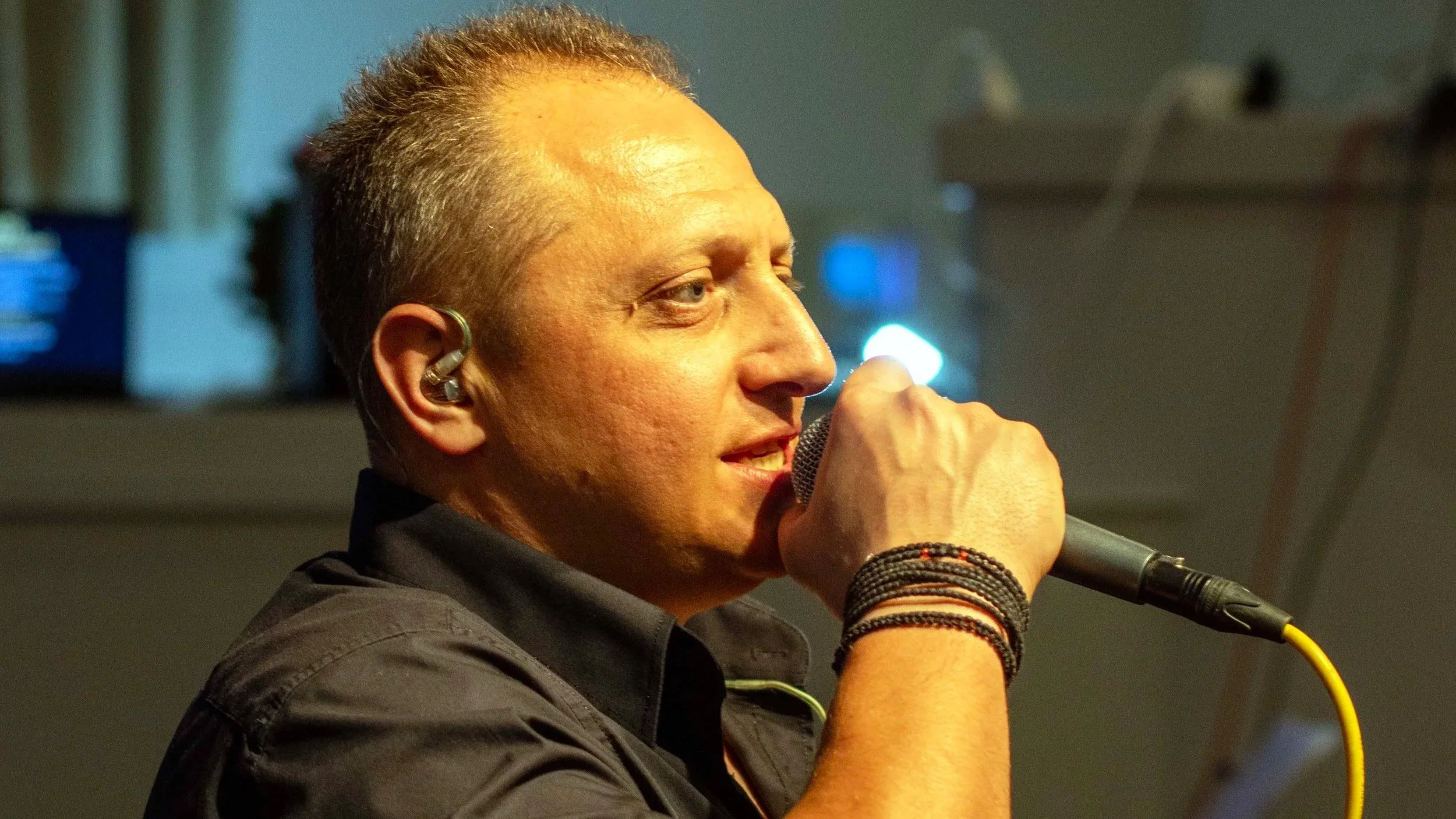 A man singing into a microphone, wearing an in-ear monitor and a dark shirt, with bracelets on his wrist.