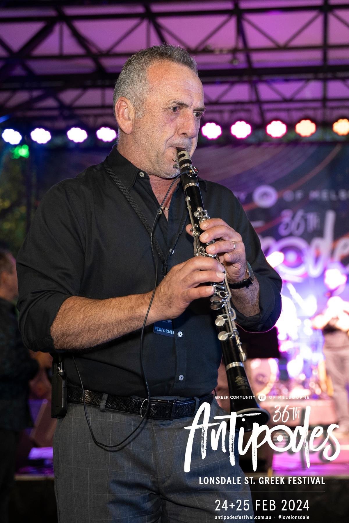 A man in a black shirt playing a clarinet at the Lonsdale St. Greek Festival, held on February 24-25, 2024, with colorful stage lights and a festival banner in the background.