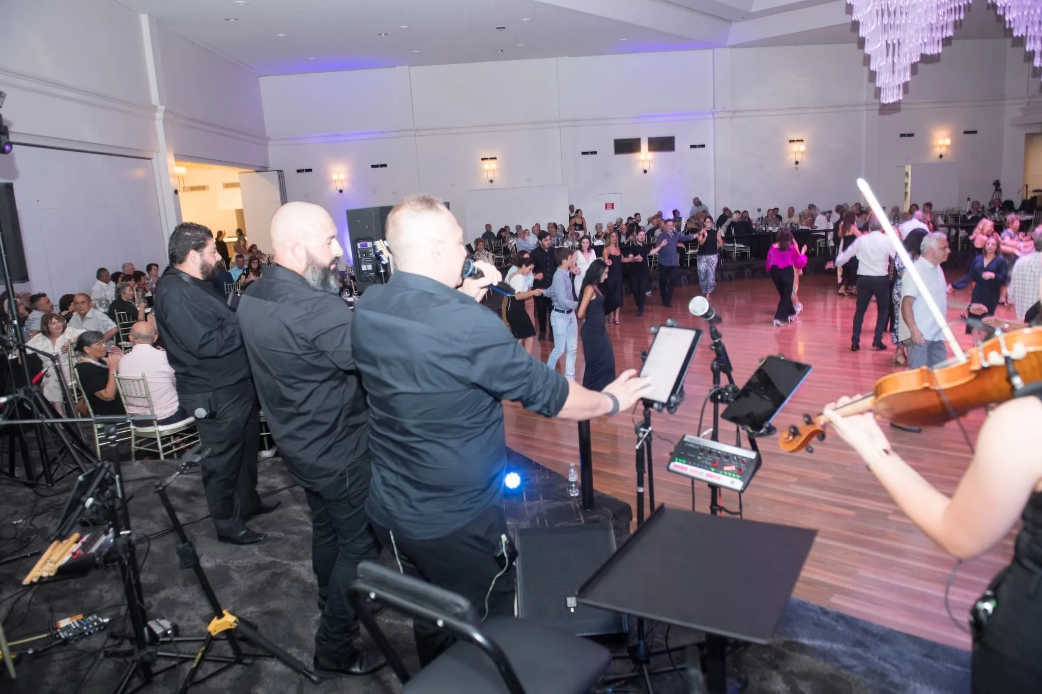 A band of musicians performs on stage with a singer, a violinist, and a microphone, while a large crowd dances on the floor in a spacious event hall with chandeliers and purple lighting.