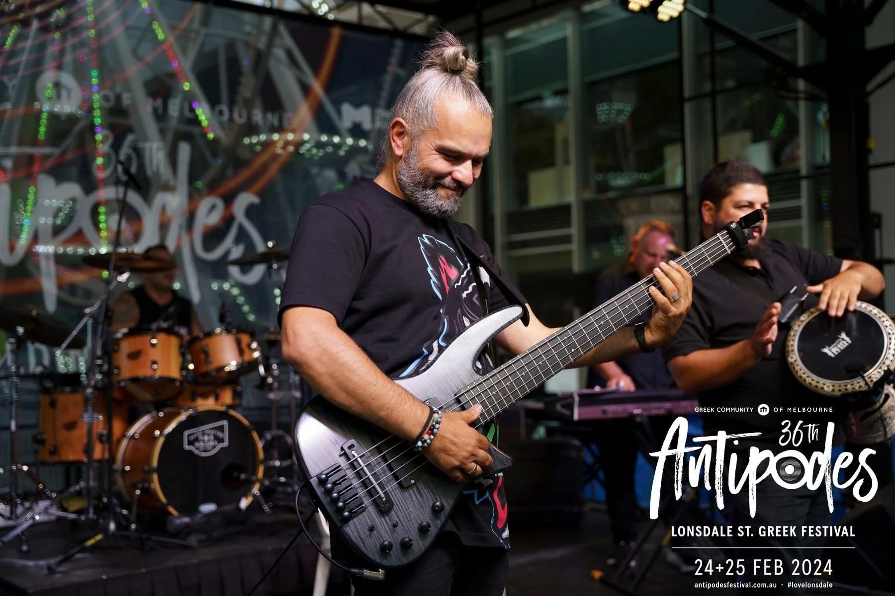 A man playing an electric guitar during a live performance at the Lonsdale St. Greek Festival, with a drummer and a percussionist in the background, at night with festival signage and lighting.