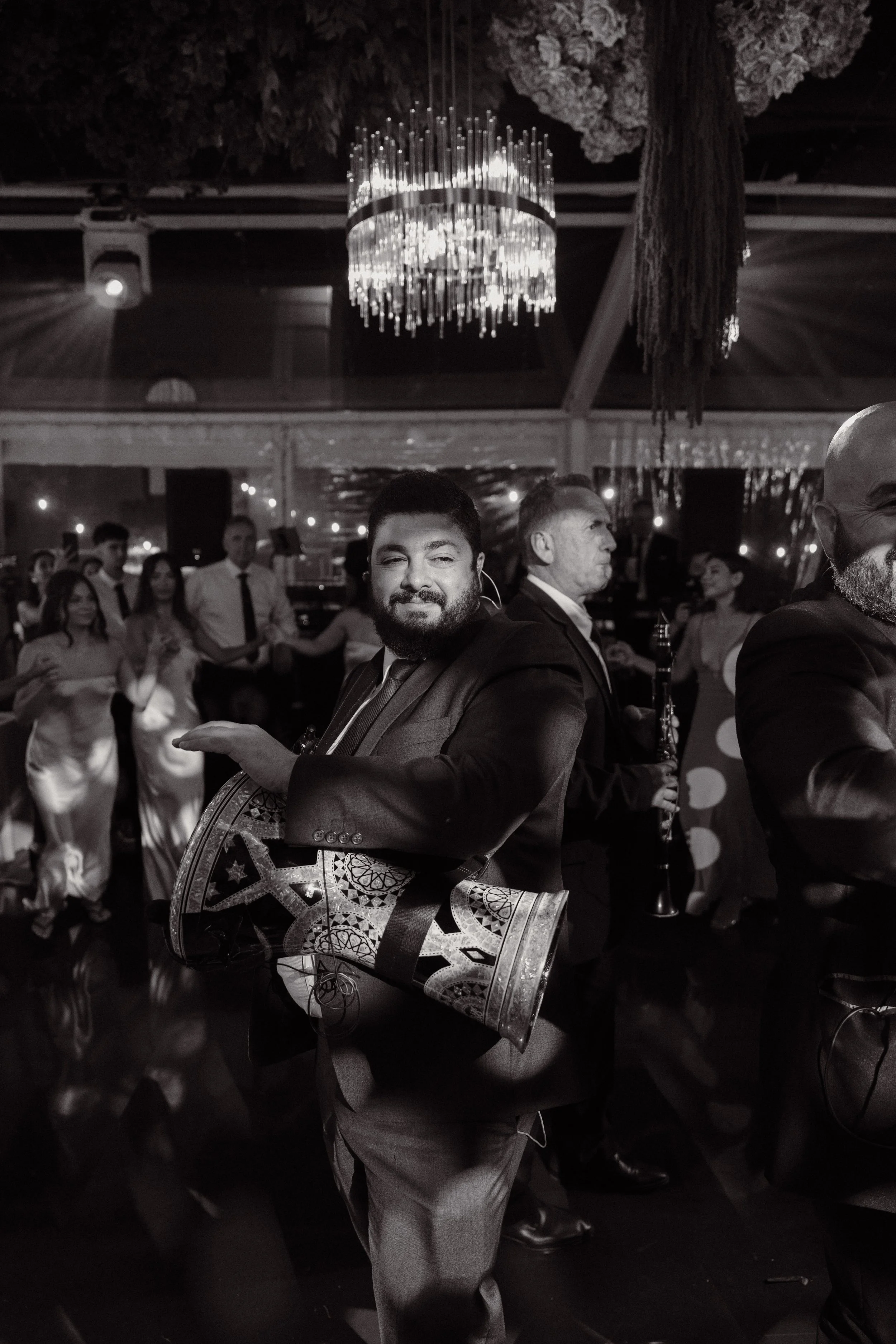 A man with a beard in a tuxedo holding a ceremonial drum at a celebration with people dancing and a chandelier in the background.