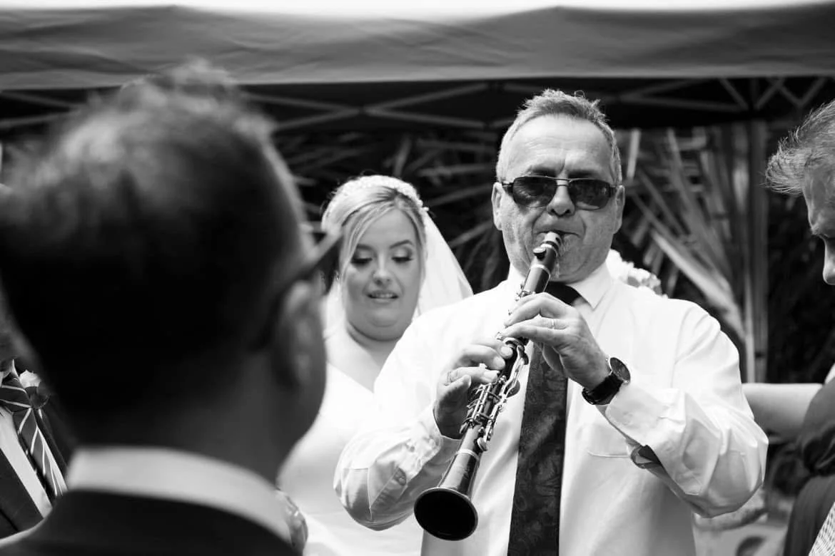 Man wearing sunglasses and a tie playing a clarinet at a wedding surrounded by guests, including a bride in a white dress and veil.