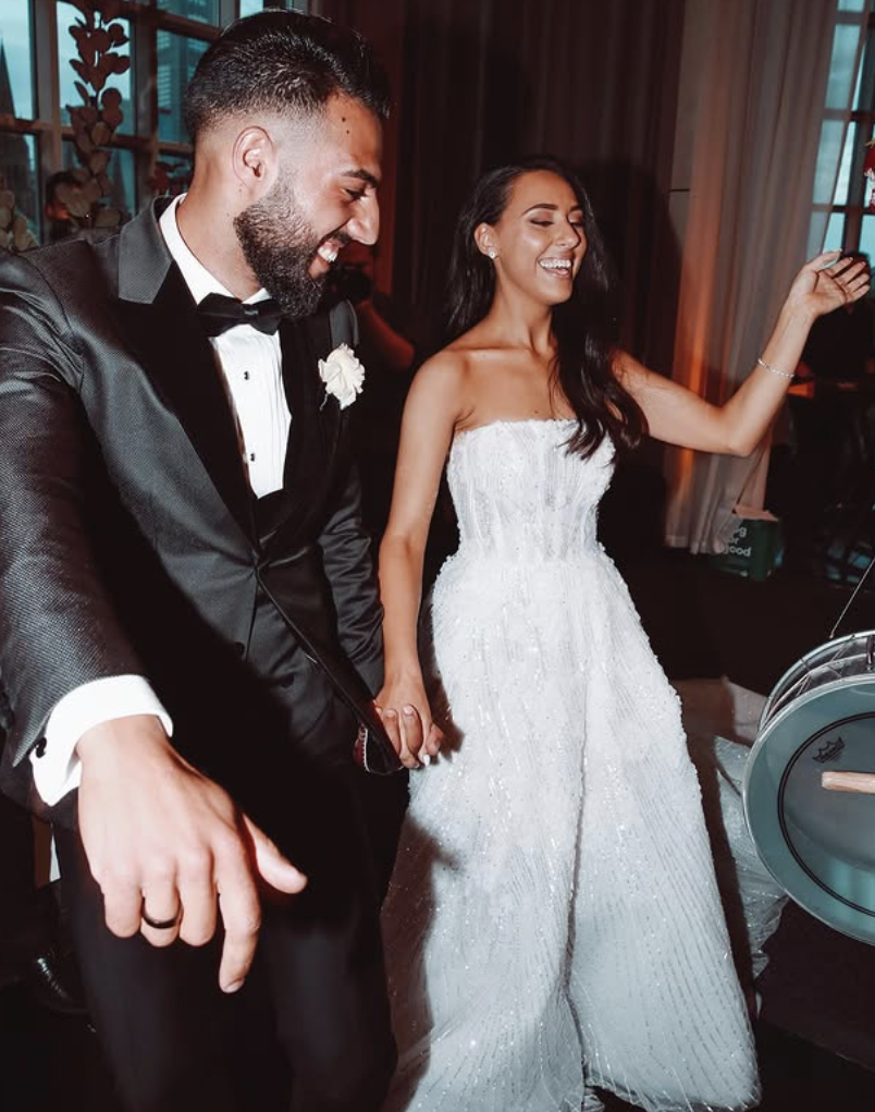 A bride and groom dancing and smiling at a wedding reception. The groom is wearing a black tuxedo with a white shirt and a black bow tie. The bride is wearing a strapless white wedding gown with beaded embellishments, and she has long dark hair. They are holding hands and appear to be enjoying the moment.