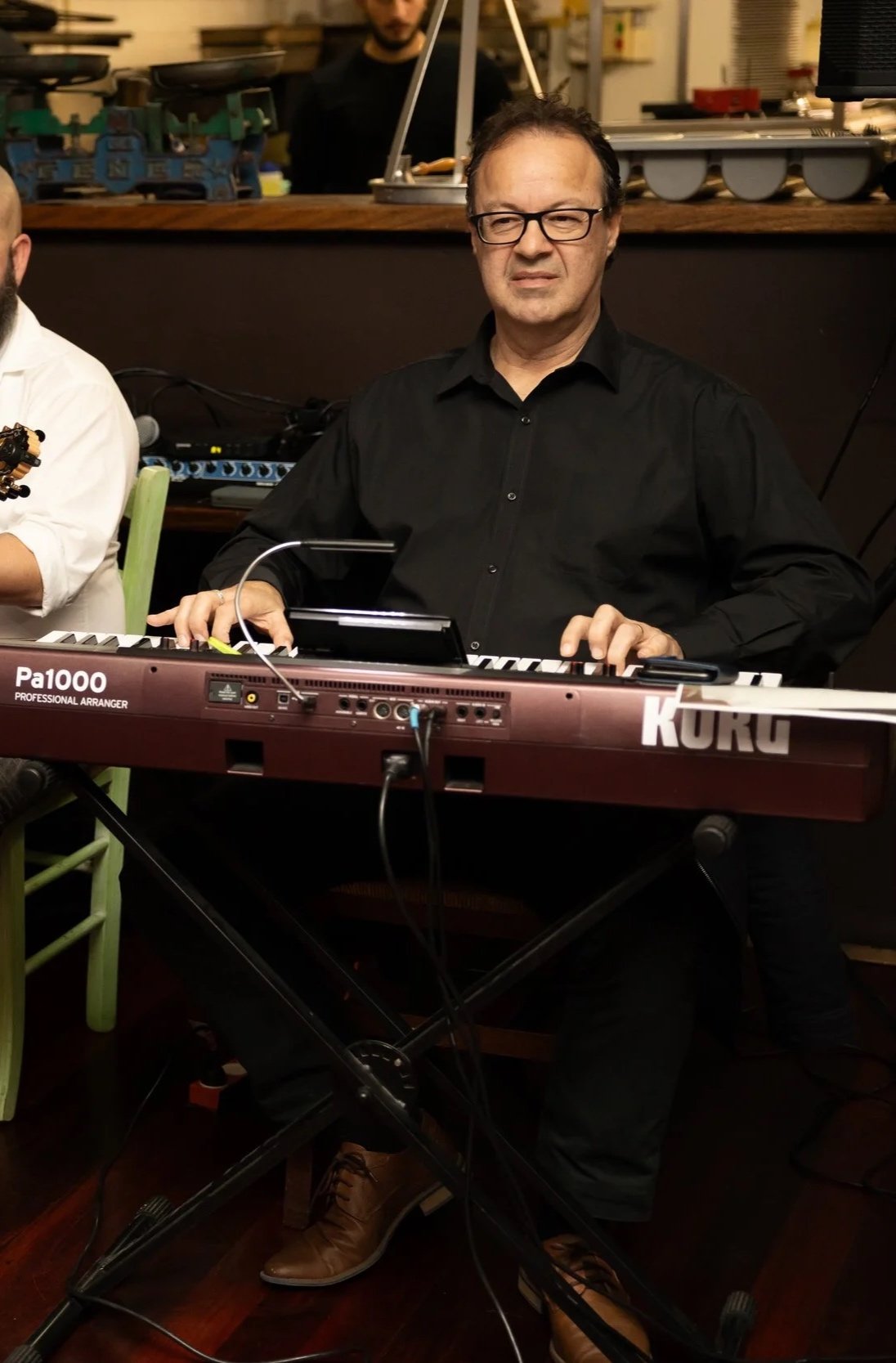 A man wearing glasses and a black shirt playing a Korg keyboard during a performance in an indoor venue.