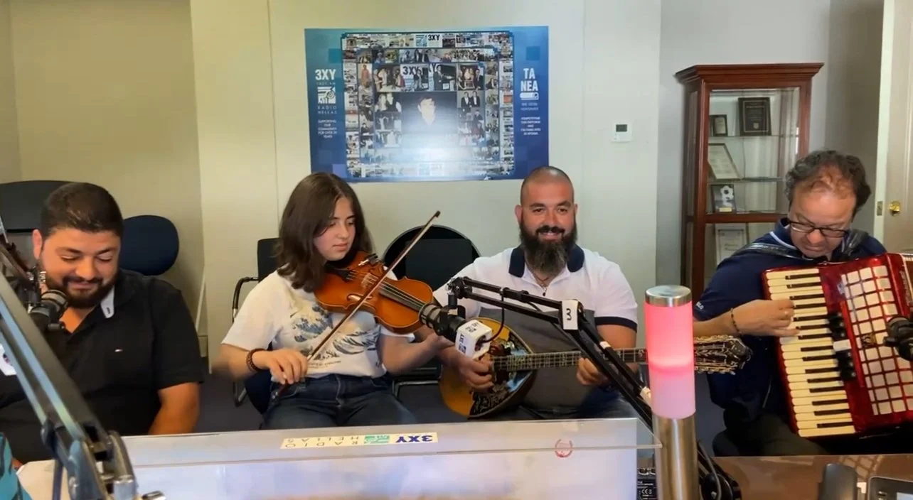 Four people playing musical instruments in a room with microphones and a framed photo collage on the wall.