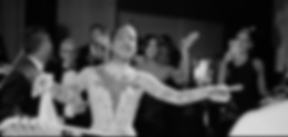 Blurry black-and-white photo of a woman smiling and raising her arms at a social gathering with several other people around her.