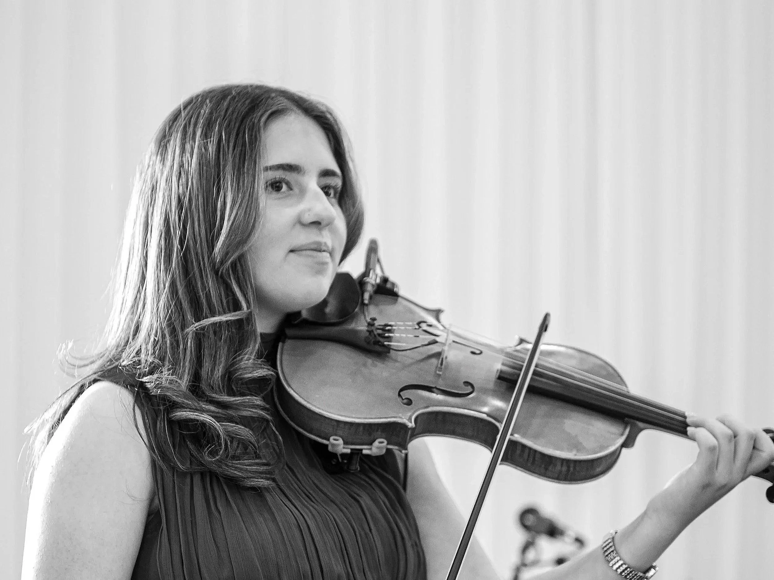 A woman with long wavy hair plays a violin, wearing a sleeveless dark top, in front of a plain background.