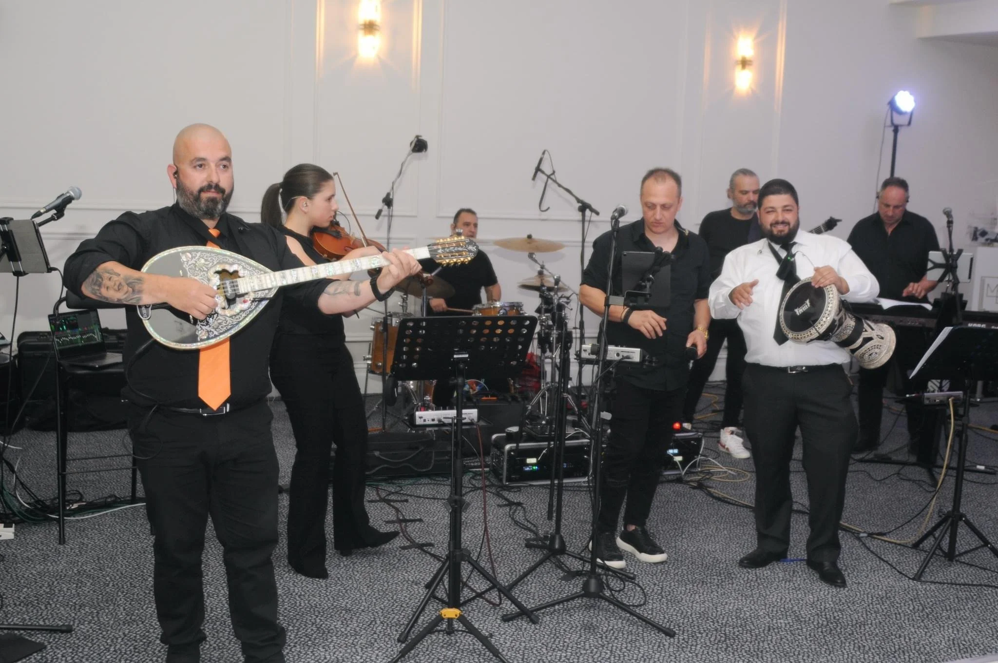 Musicians performing at an indoor event with instruments including a guitar, violin, drums, and hand drums, with microphones and sound equipment.