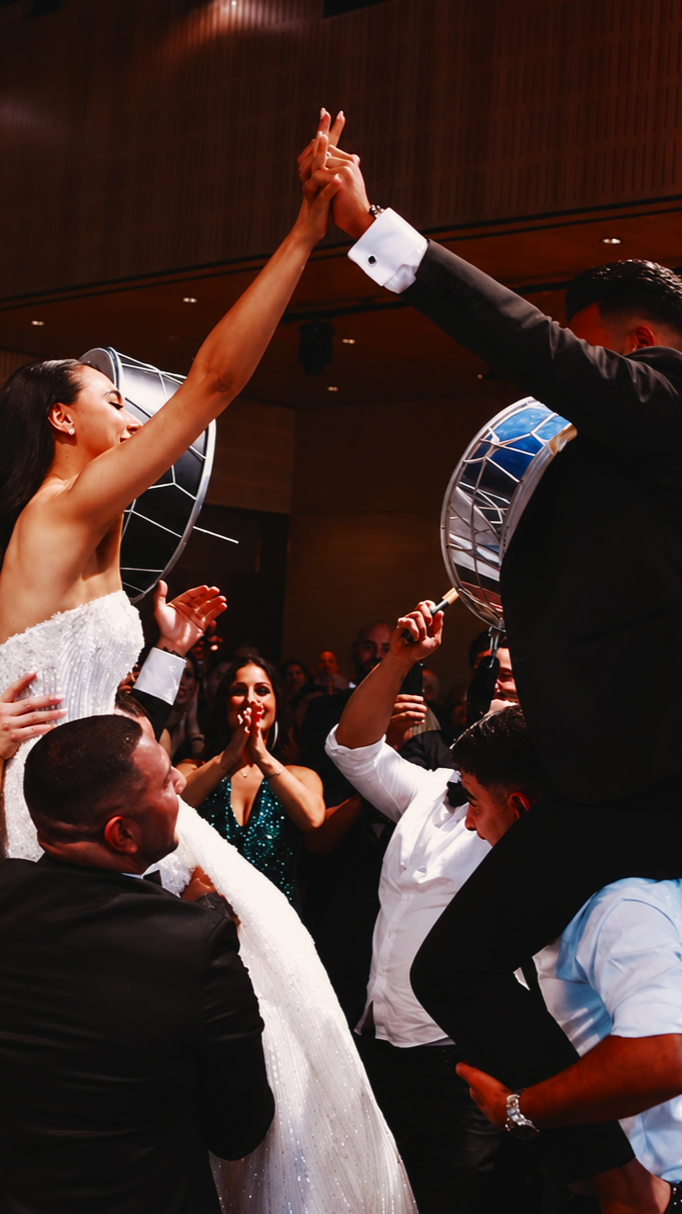 Couple dancing during a celebration, the woman in a white dress and the man in a black suit, surrounded by guests clapping and smiling.