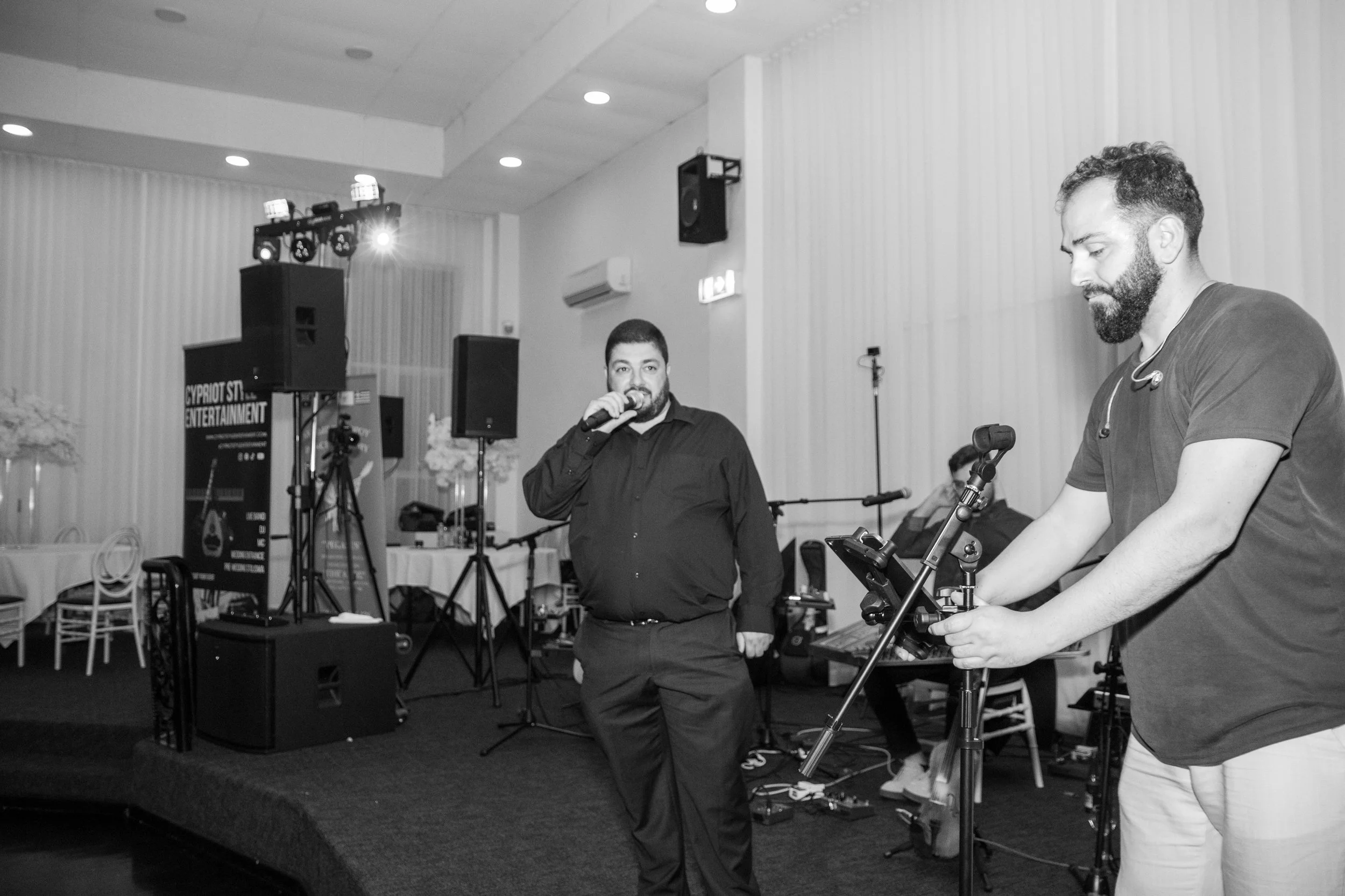 Two men are setting up or preparing equipment on a stage at an indoor event or performance. One man is speaking into a microphone, and the other man is handling equipment, possibly a music or DJ controller. The background features audio and lighting equipment, including speakers and a lighting rig.