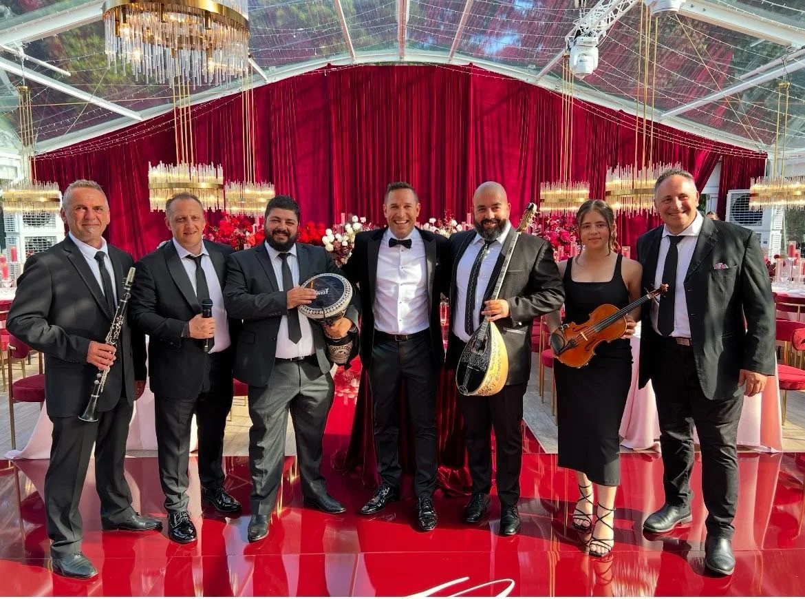 Music band dressed in formal black suits and a female violinist in black dress, standing in a decorated event hall with red drapes, chandeliers, and tables, posing for a photo.