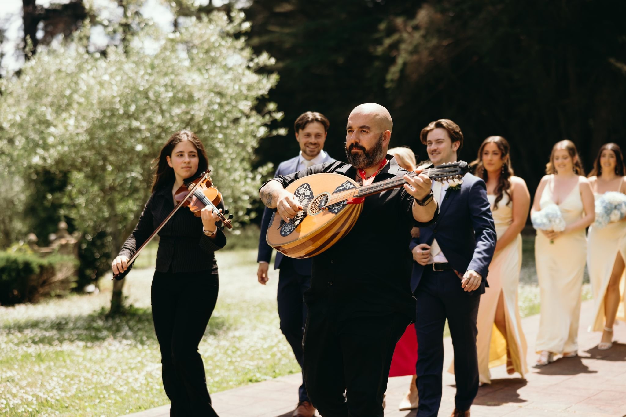 A musician playing a guitar in a wedding procession surrounded by bridesmaids and groomsmen outdoors.