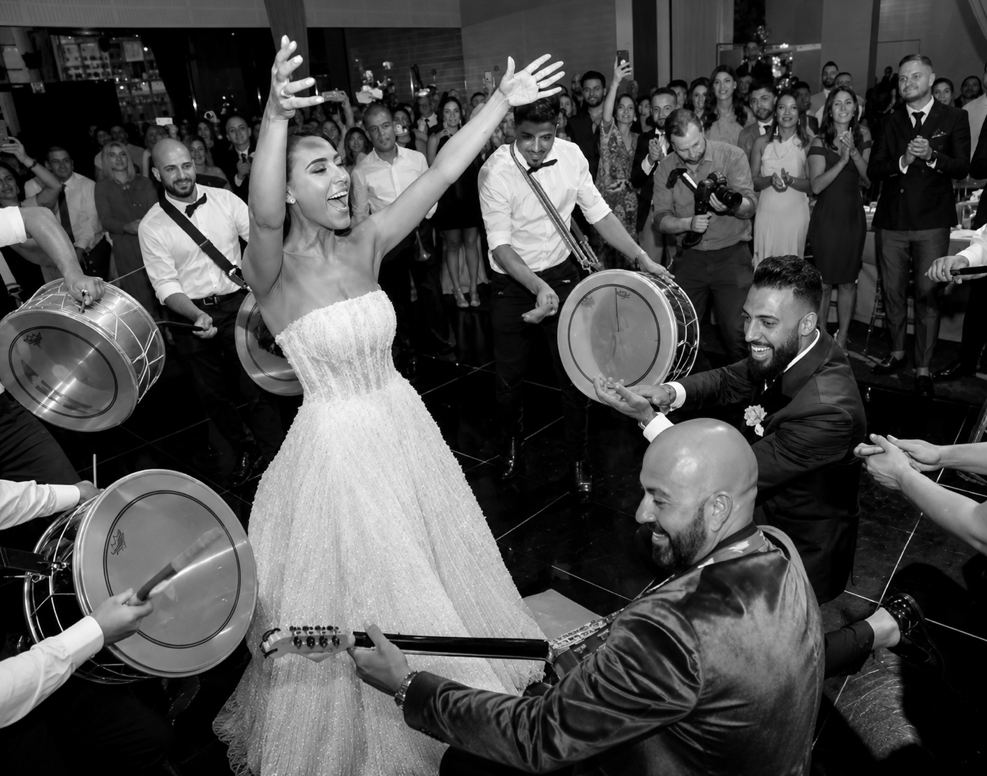 A bride in a strapless wedding dress is dancing with male musicians playing drums and guitar, surrounded by guests at a wedding reception.