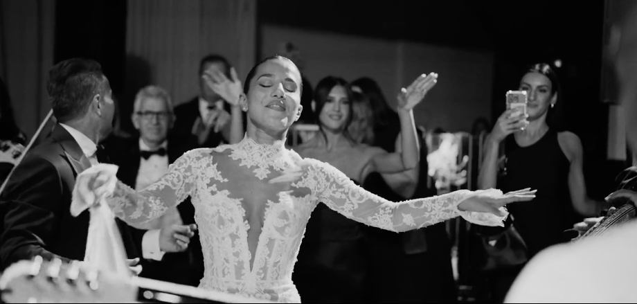 A woman in a lace dress dances with her arms outstretched at a celebration or party, surrounded by other people, some taking photos, in a festive environment.