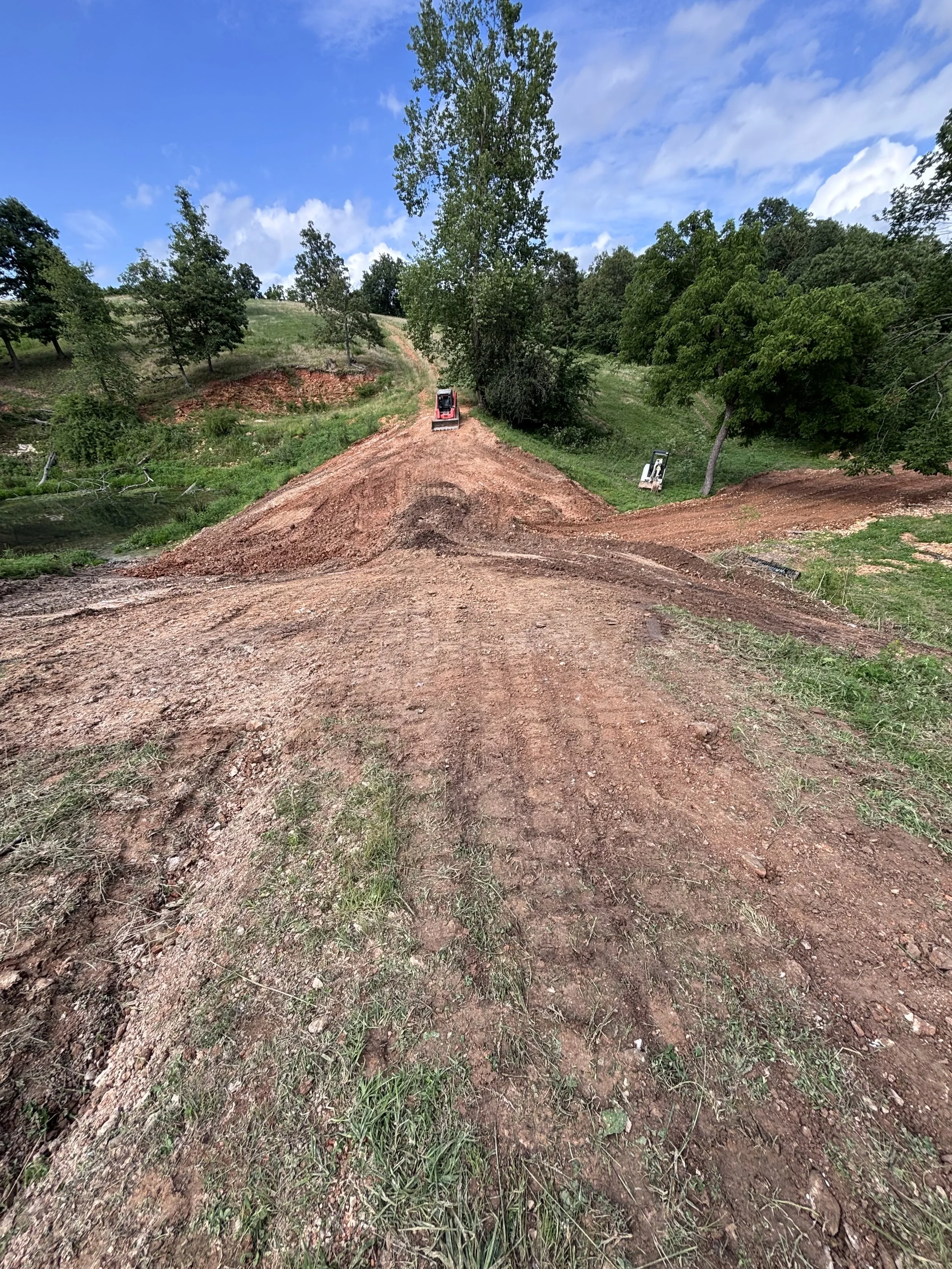 Dirt trail cut into a hillside with construction vehicles in the background under a partly cloudy sky.