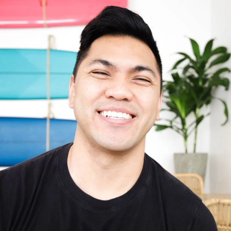 Smiling young man with black hair in a casual setting with colorful surfboards and a potted plant in the background.