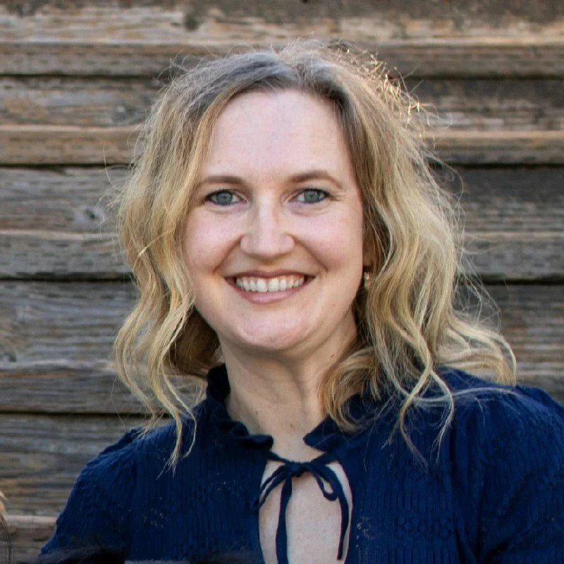 A woman with blonde, wavy hair smiling in front of a wooden background.