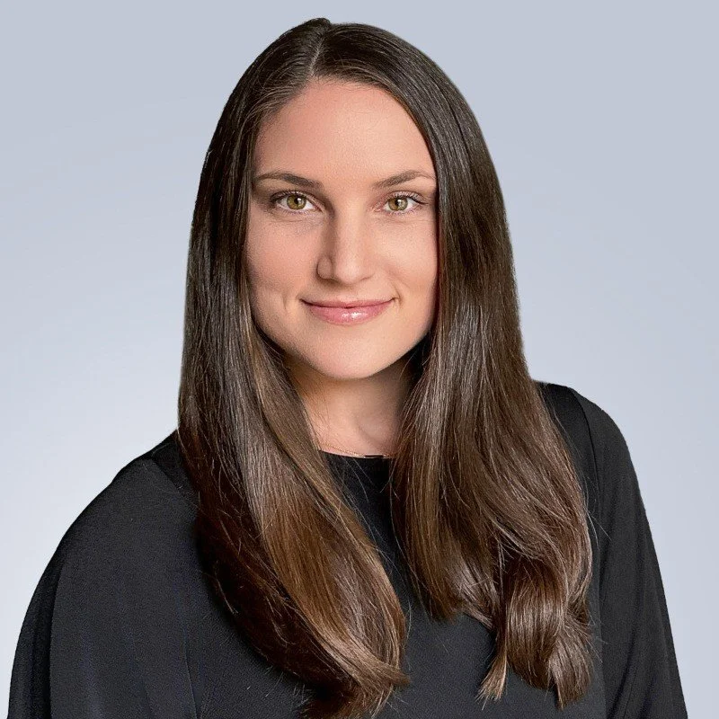A woman with long brown hair and hazel eyes, smiling, wearing a black top, against a light gray background.