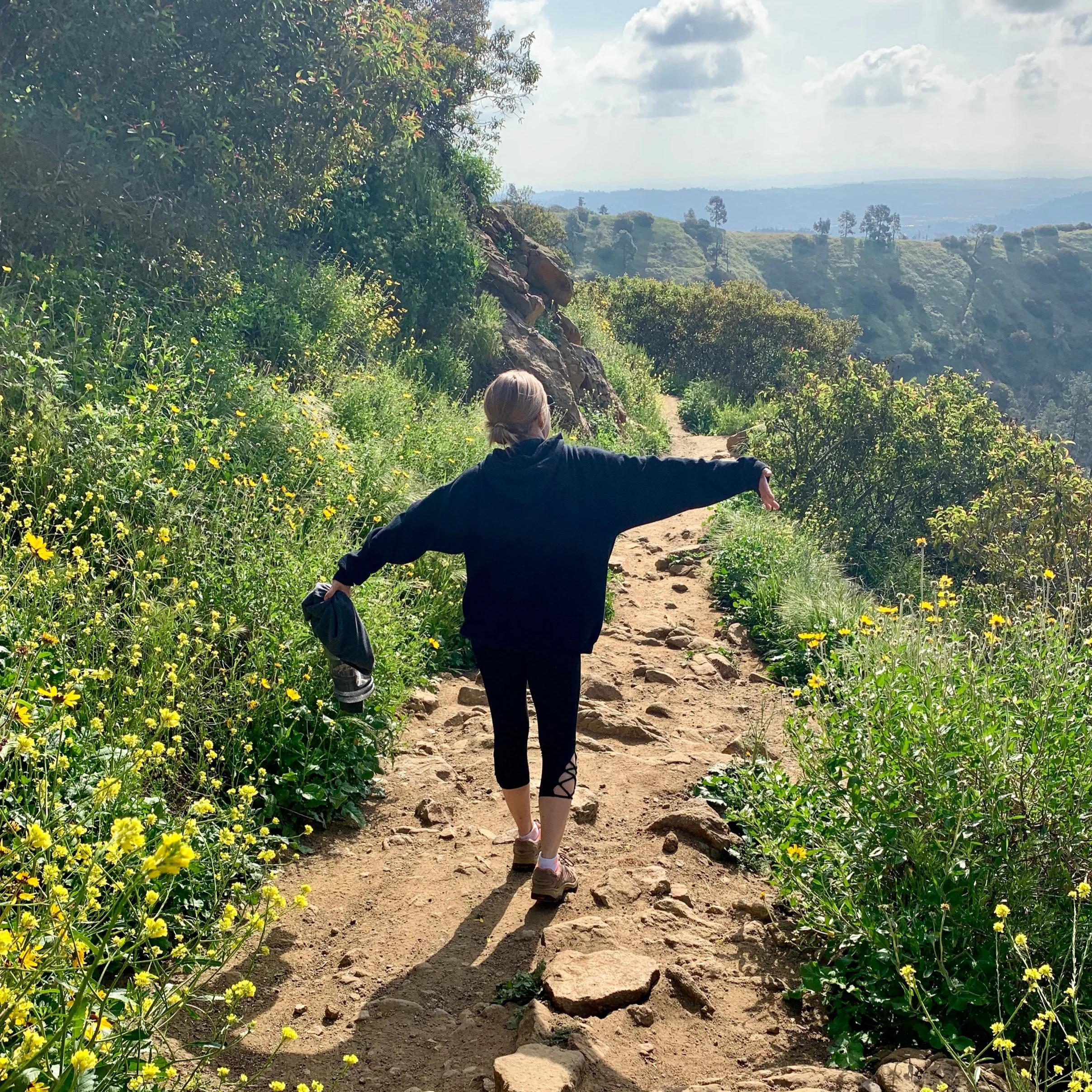 Person hiking on a dirt trail surrounded by green plants and yellow flowers, with scenic hills and cloudy sky in the background.