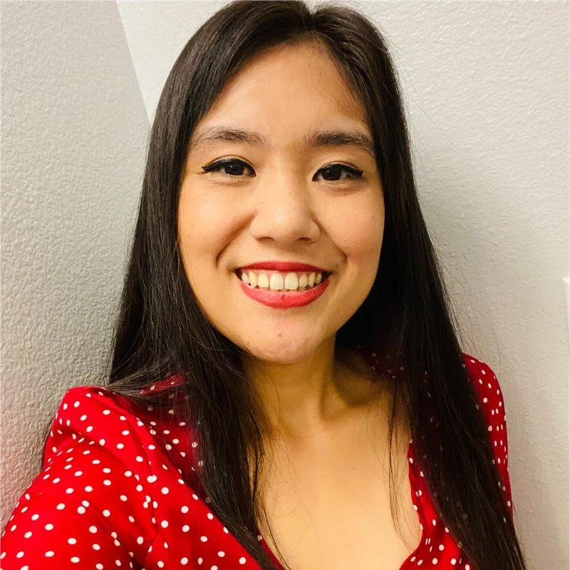 A woman with long black hair and a bright smile, wearing a red dress with white polka dots, standing against a plain wall.