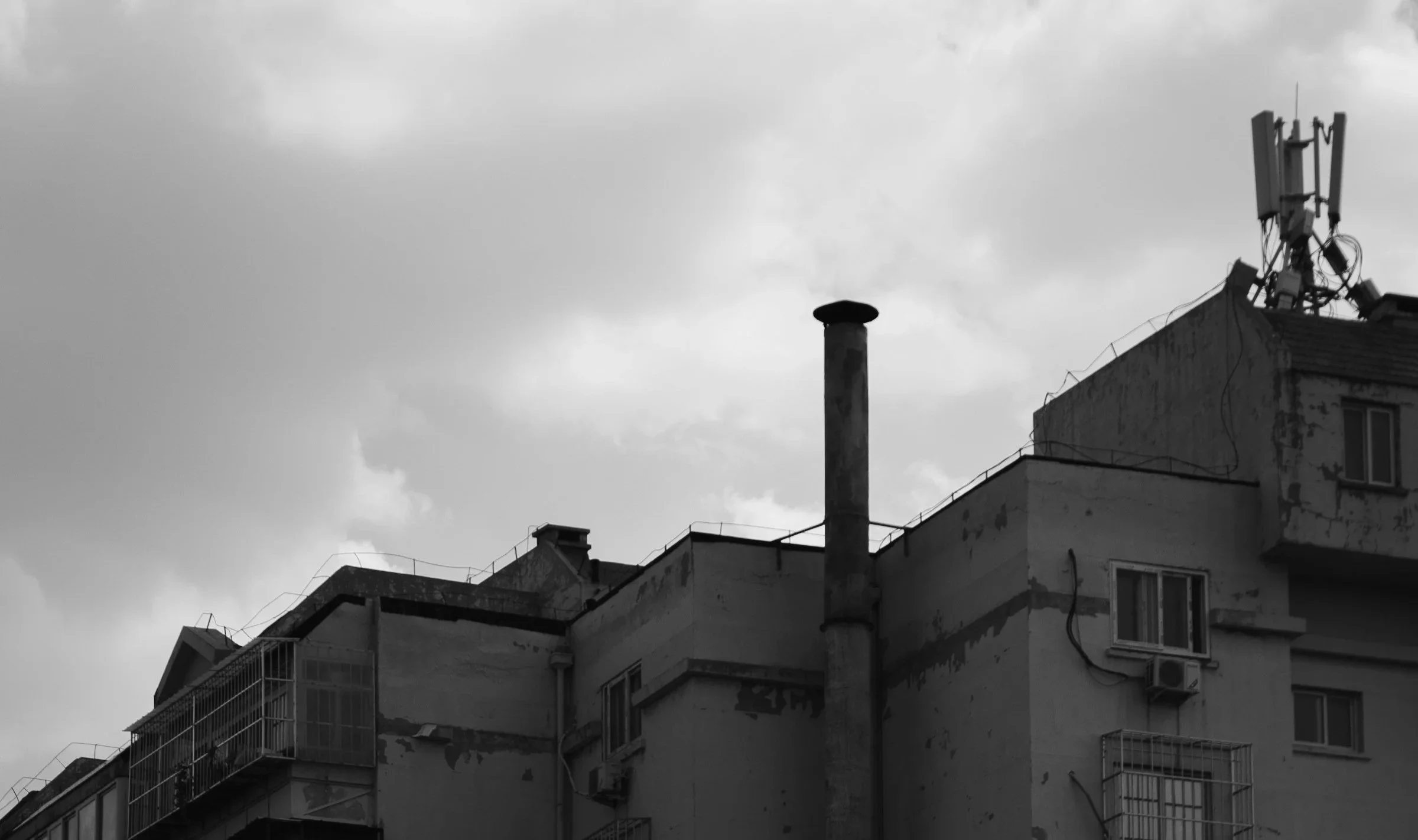 Black and white photo of an apartment building with a chimney and a cell tower on the roof under cloudy skies, appearing like a silhouette of a growth chart.