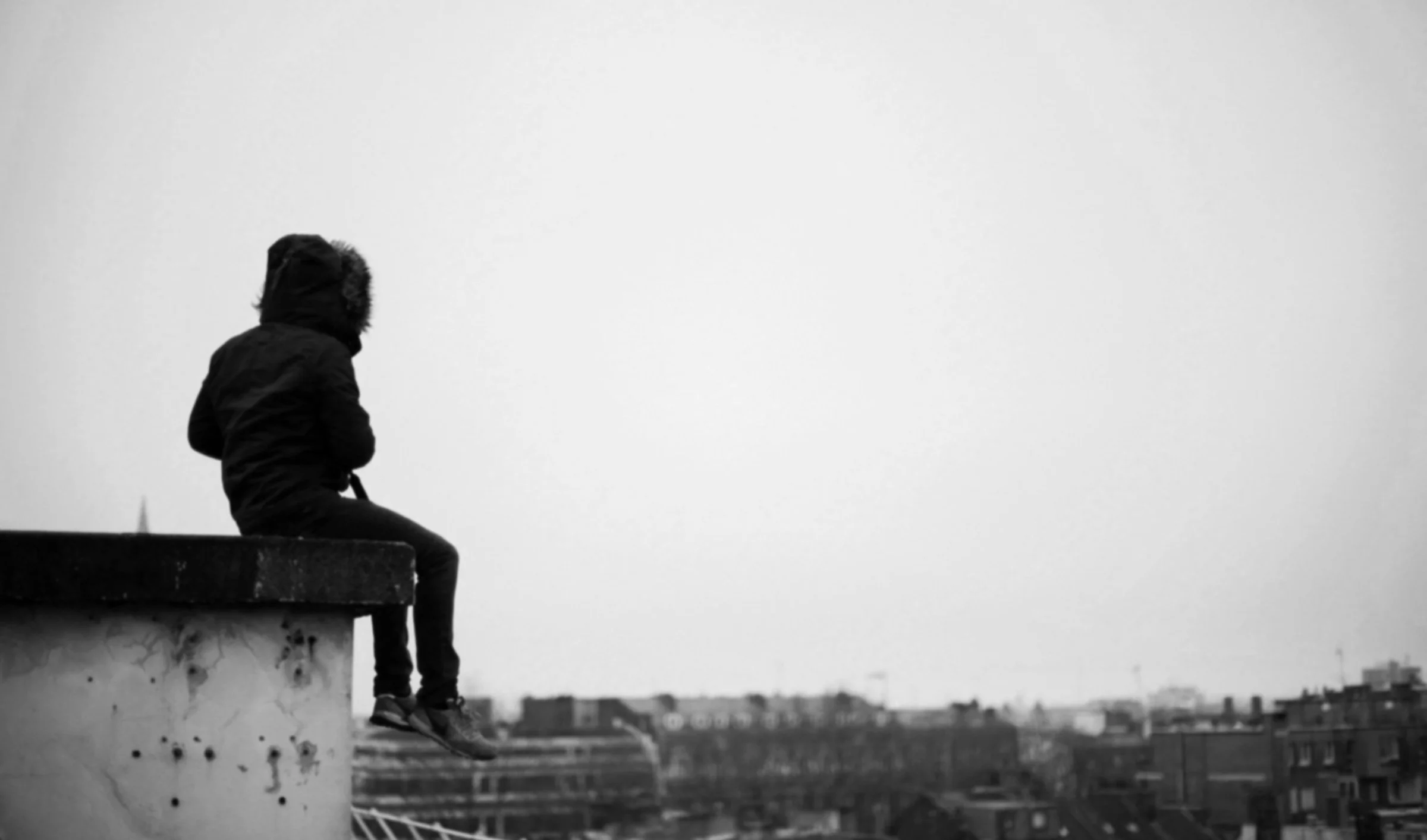 A person sitting alone on a rooftop ledge, wearing a hooded jacket, overlooking a cityscape in black and white, representing reaching the top and growth.