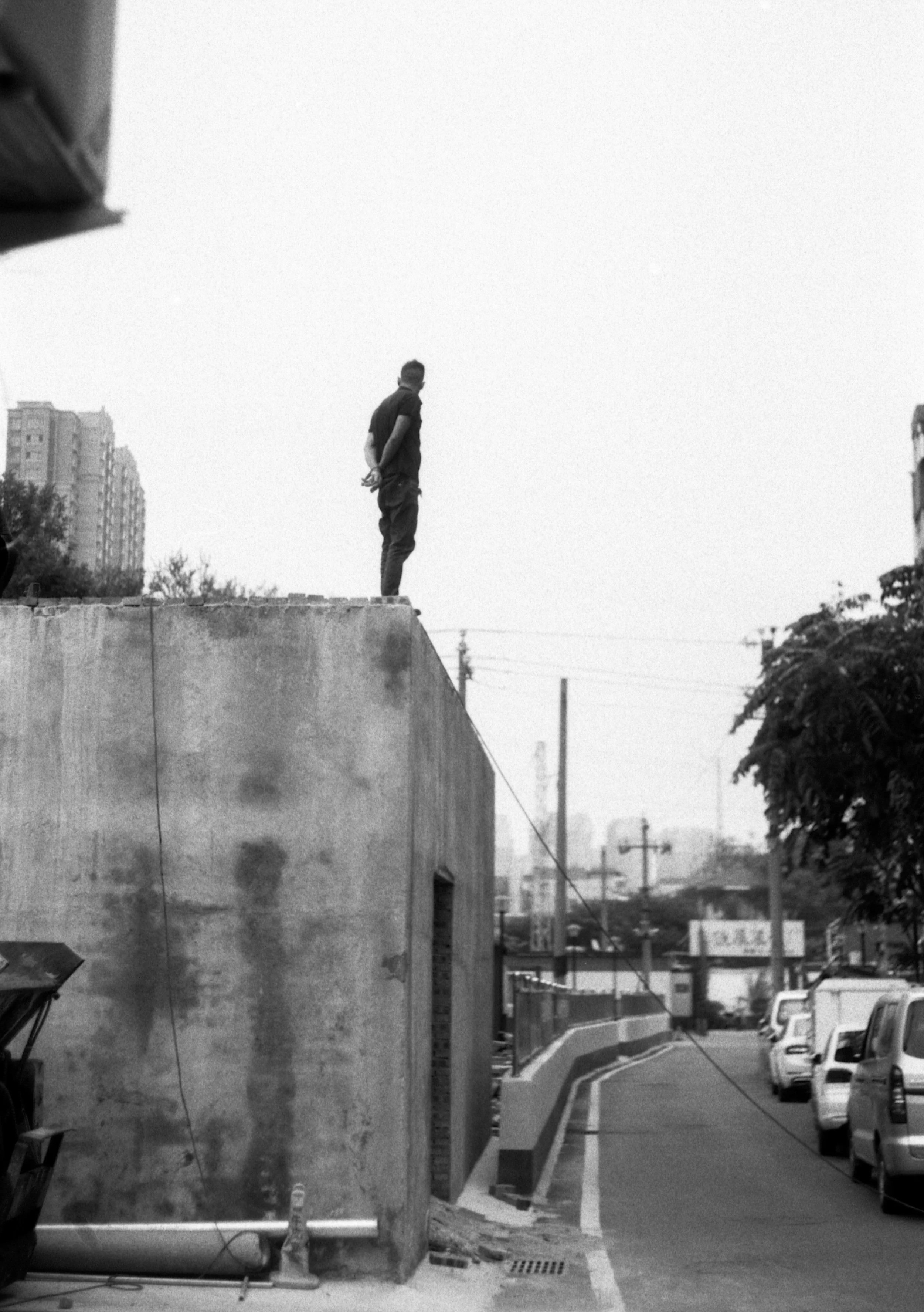 A man standing on the edge of a rooftop, looking out over an urban street scene in black and white, representing reaching the top and growth.
