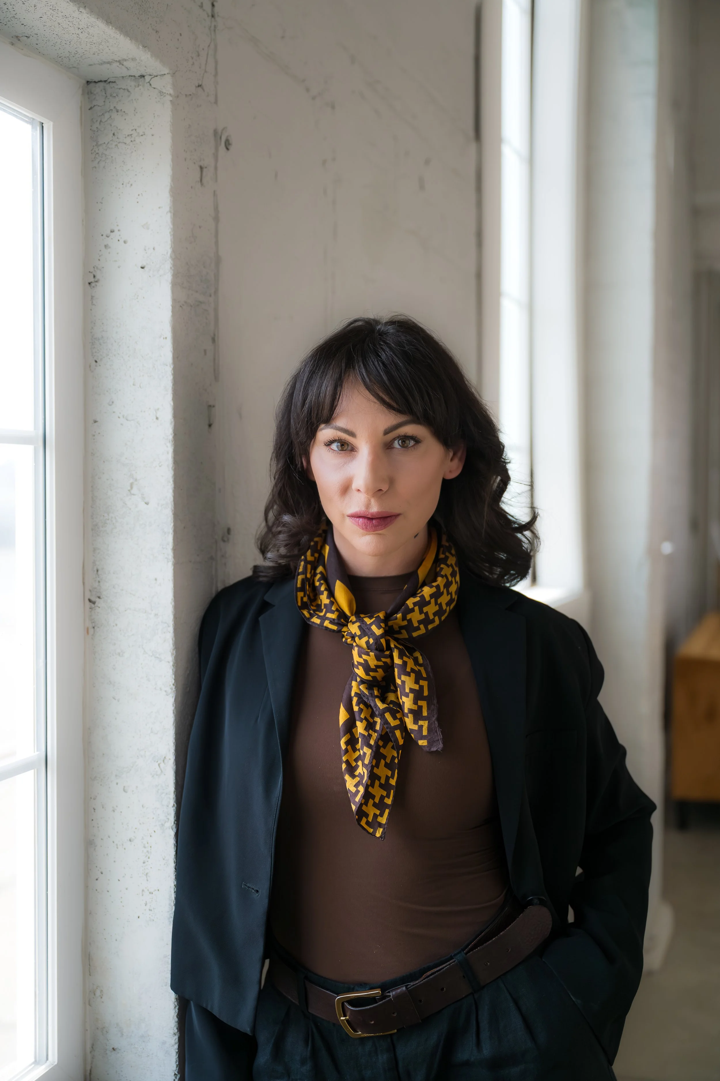 A woman with shoulder-length dark hair wearing a black blazer, brown top, and a yellow and brown patterned scarf tied around her neck, standing indoors near a window with a neutral expression.