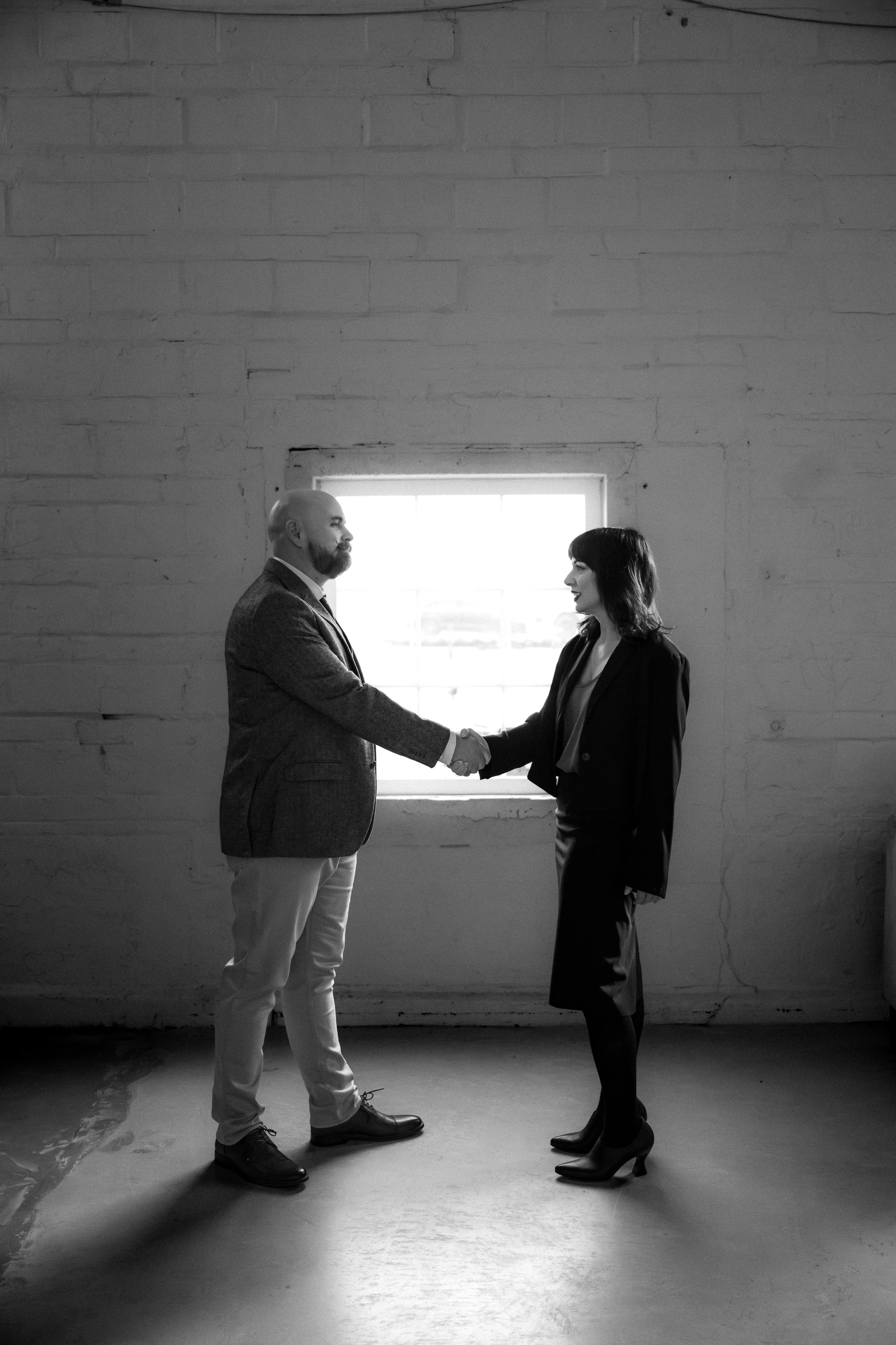 Two people shaking hands in an industrial-style room with a window in the background, black and white photograph.